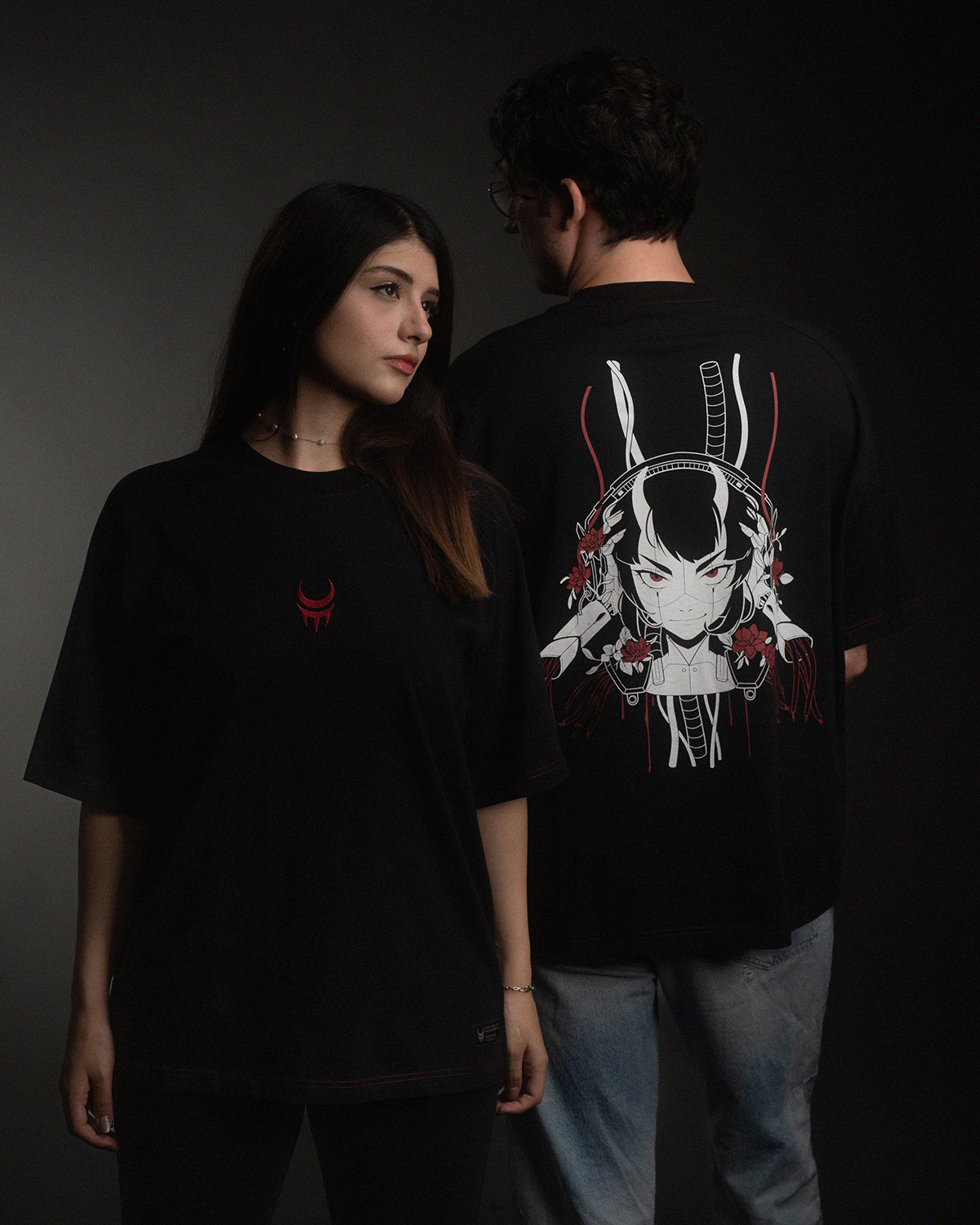 Zwei Personen tragen schwarze T-Shirts. Die Vorderseite zeigt ein kleines rotes Mondkrallen-Symbol auf der Brust, die Rückseite ein großflächiges Printmotiv mit einer stilisierten Cyborg-Figur mit Hörnern, Blumen und Kabeln in Weiß, Rot und Grau.