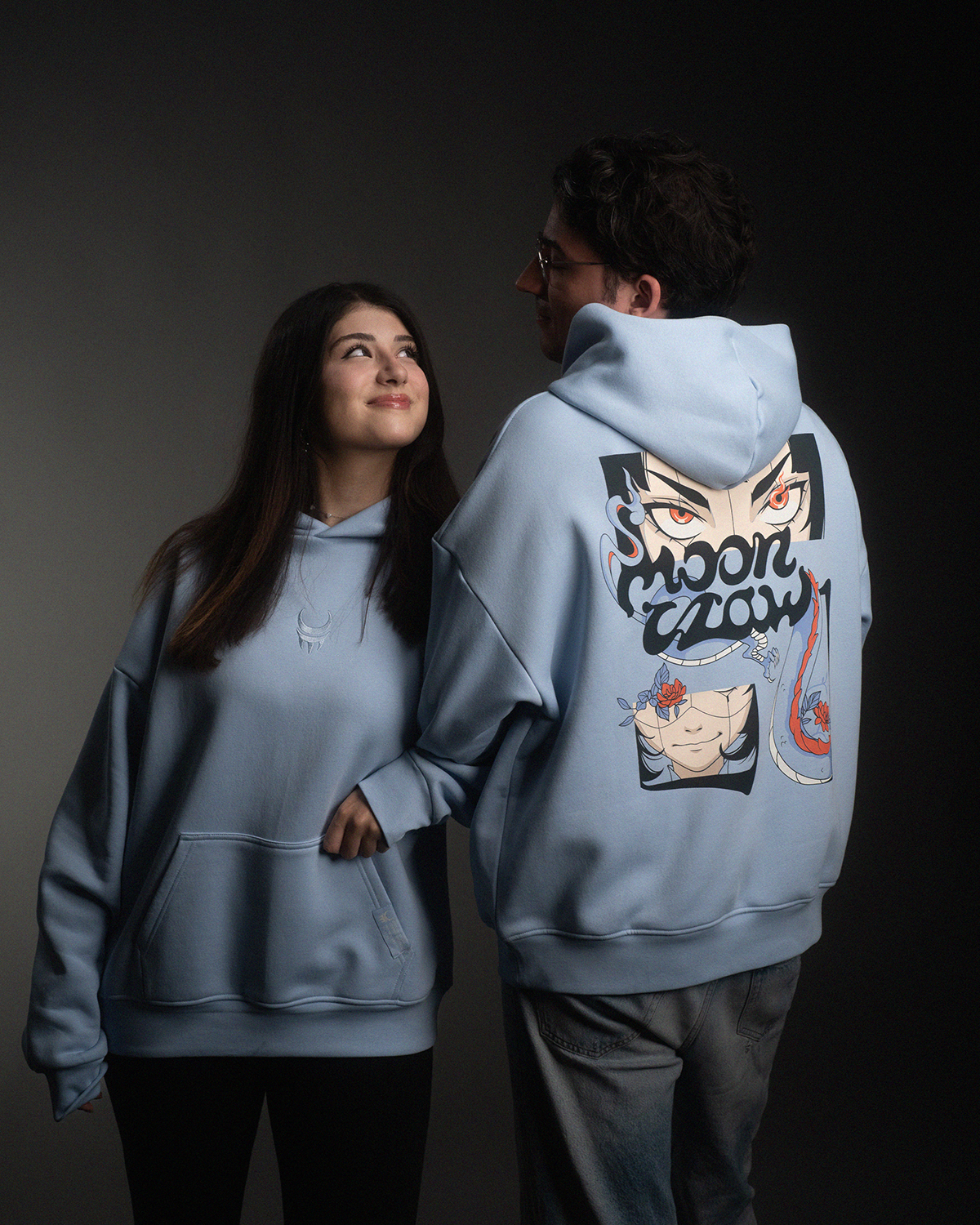 Zwei Personen tragen hellblaue Oversized-Hoodies. Der linke Hoodie zeigt ein gesticktes Logo auf der Brust, der rechte ein großflächiges Rückendesign mit einem illustrierten Gesicht, dem Schriftzug „moon claw“ und grafischen Elementen in Rot, Schwarz und Blau.