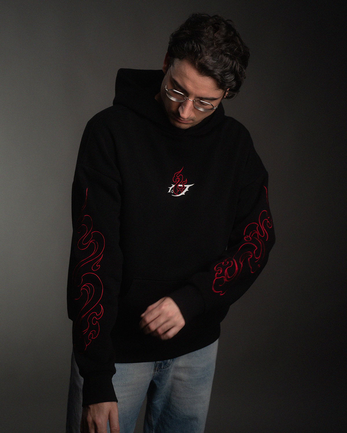 Schwarzer Hoodie mit roter Flammenstickerei auf der Brust und beidseitiger Stickerei an den Ärmeln. Getragen von einem Model mit Brille, Blick nach unten gerichtet.