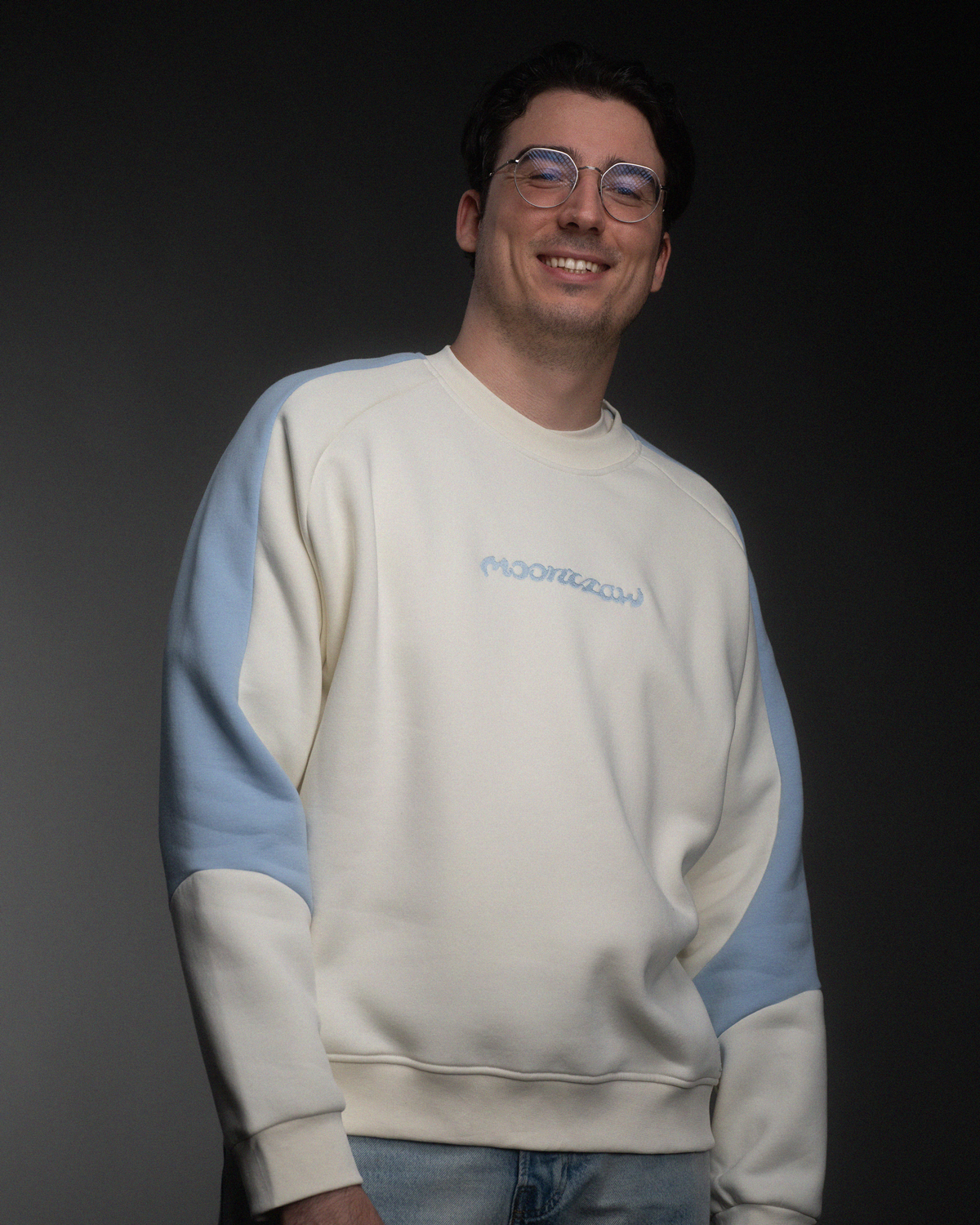 Ein Mann trägt ein cremefarbenes Sweatshirt mit hellblauen Ärmeln. Auf der Brust ist in hellblauer Stickerei das Wort „moonclaw“ im geschwungenen Schriftzug platziert.