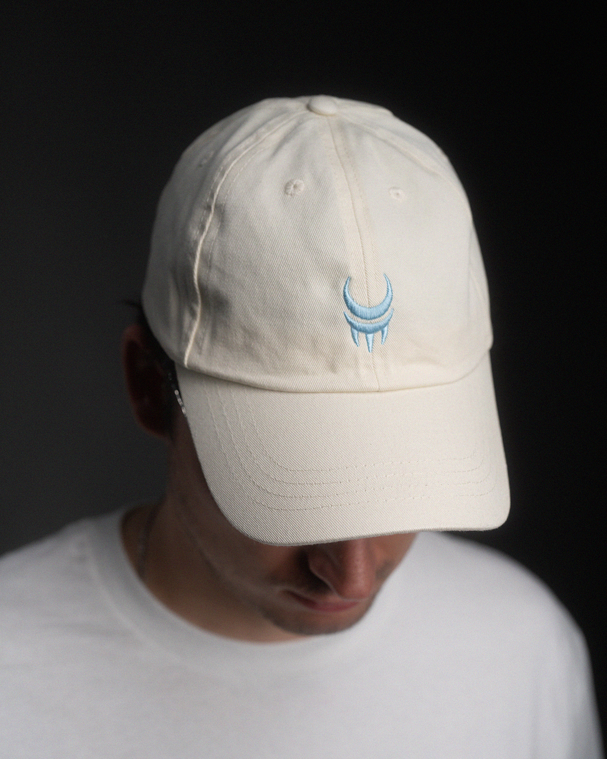 Cremefarbene Baseball-Cap mit hellblauem Stickmotiv aus Sichel und Krallen auf der Front, getragen von einer Person mit gesenktem Blick.