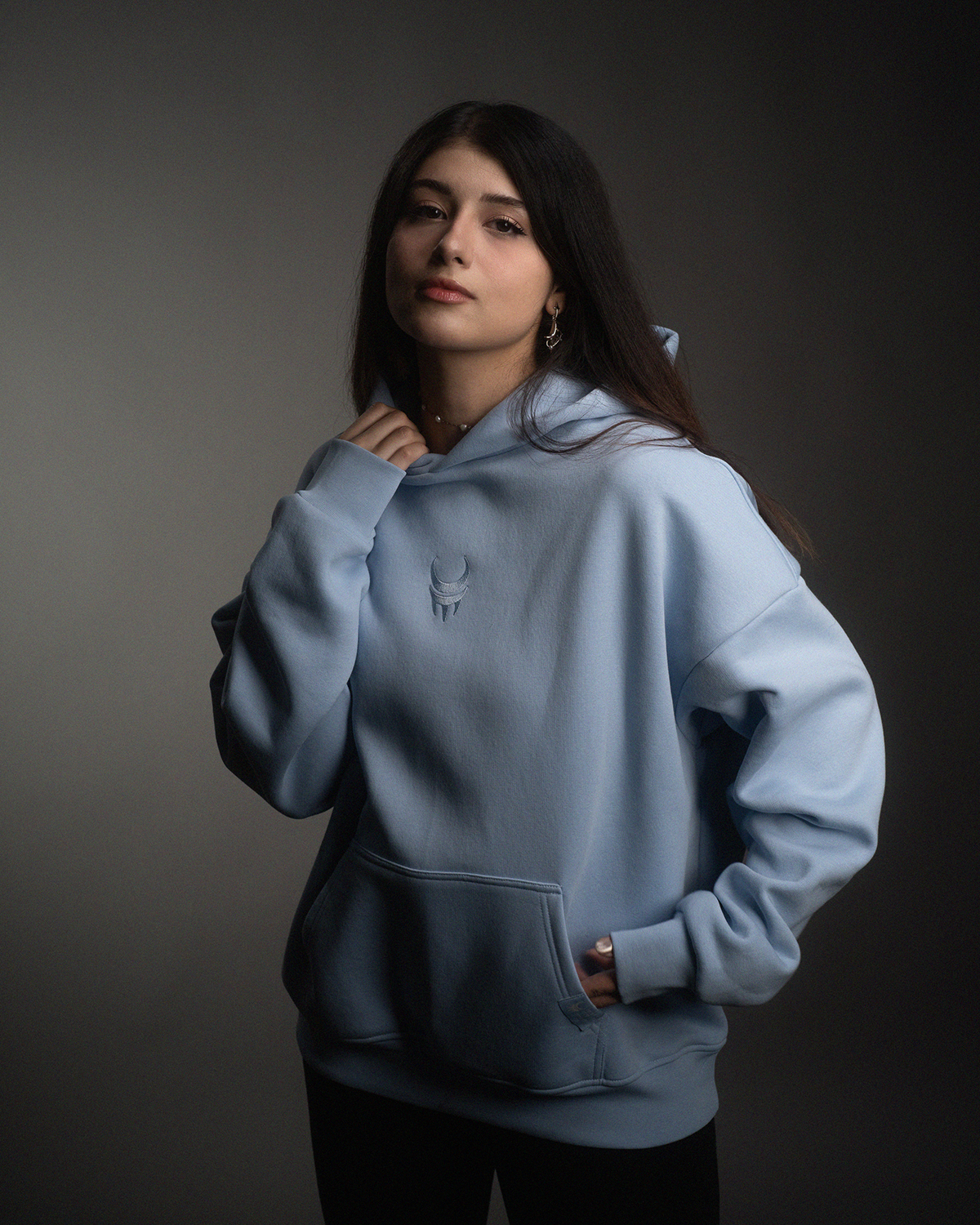 . Frontansicht eines hellblauen Hoodies mit weißem gesticktem „moon claw“-Symbol auf der Brust. Die Kapuze liegt locker im Nacken, das Design ist schlicht und tonal.