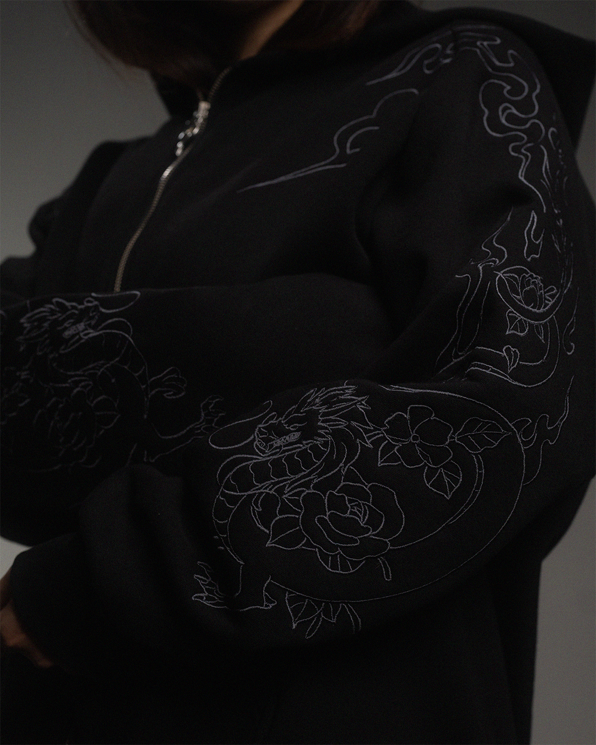 Detailansicht des linken Ärmels der schwarzen Kapuzenjacke. Sichtbar ist eine Ton-in-Ton-Stickerei mit einem Drachen- und Blumenmotiv.