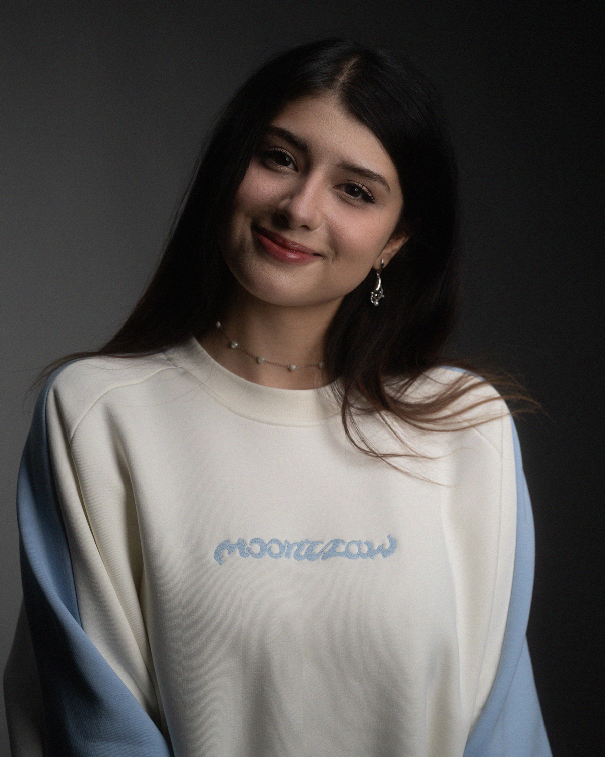 Eine Frau trägt ein cremefarbenes Sweatshirt mit hellblauen Ärmeln. Auf der Brust ist der Schriftzug „moonclaw“ in hellblauer Stickerei zu sehen.