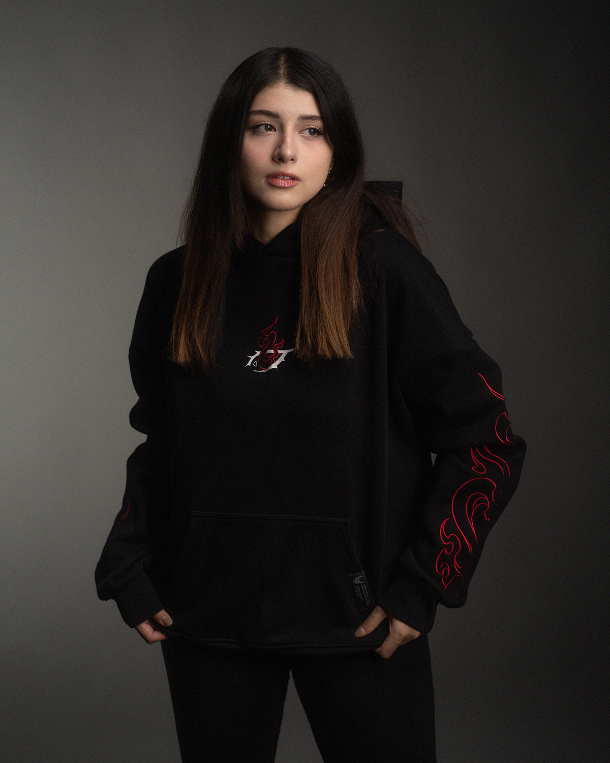 Schwarzer Hoodie mit zentralem Brustmotiv aus roter Flamme und weißem Symbol, dazu rote Flammenstickereien auf beiden Ärmeln. Getragen von einer Frau in neutraler Studio-Umgebung.