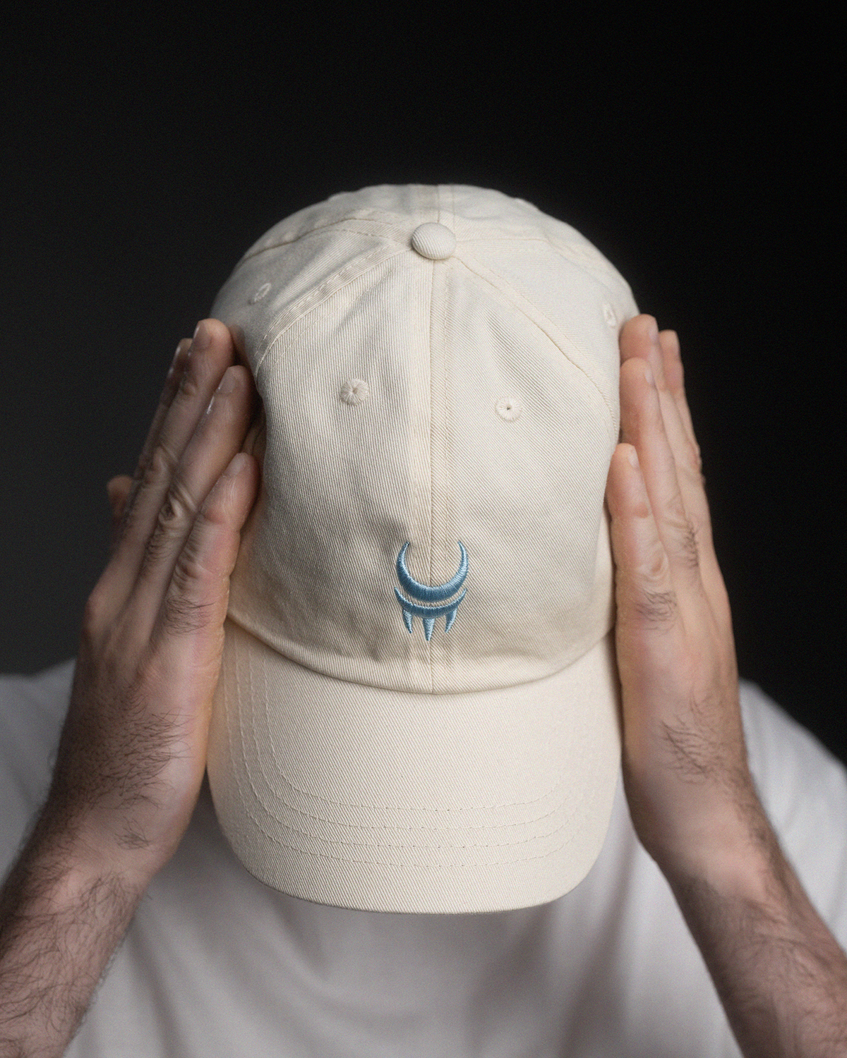 Cremefarbene Baseball-Cap mit hellblauer Sichel- und Krallenstickerei auf der Vorderseite, von oben fotografiert, getragen von einer Person mit beiden Händen seitlich am Kopf.