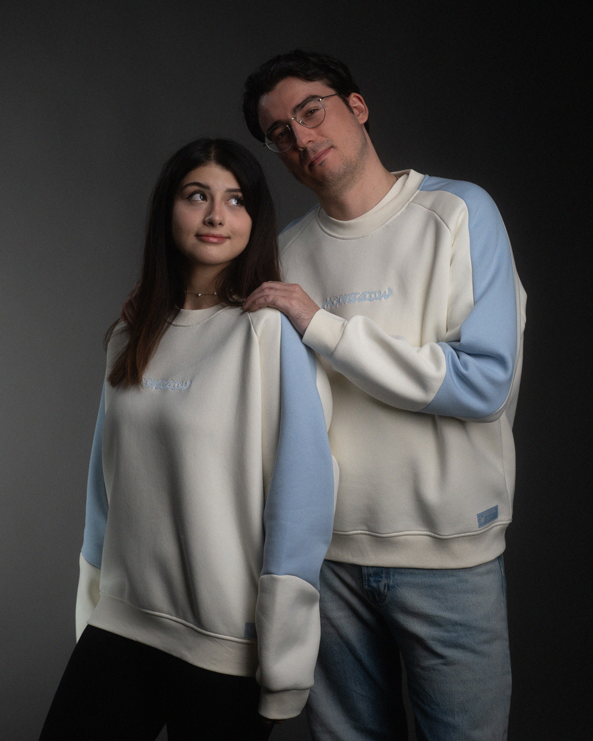 Eine Frau und ein Mann tragen identische cremefarbene Sweatshirts mit hellblauen Ärmeln. Beide Oberteile zeigen auf der Brust den gestickten Schriftzug „moonclaw“.