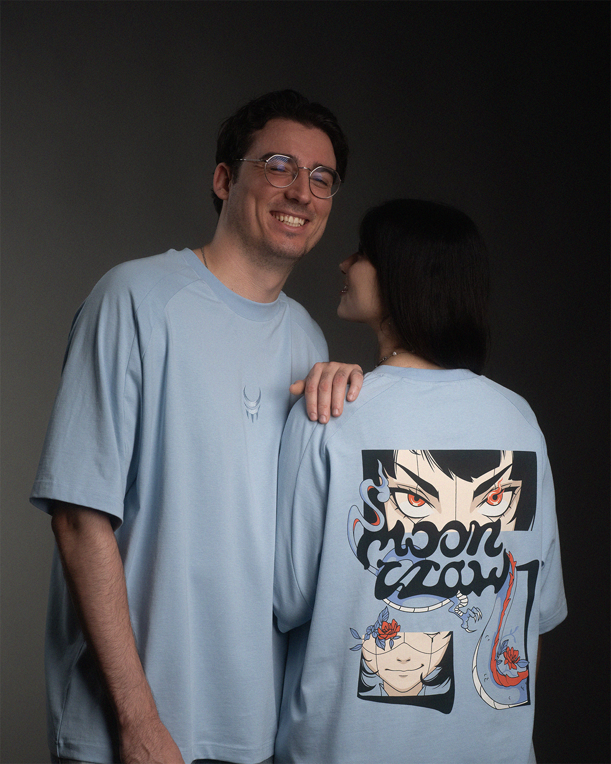 Zwei Personen mit hellblauen T-Shirts, das Rückenteil zeigt einen großflächigen Comic-Print mit Charakterillustration und „MoonClaw“-Schriftzug.