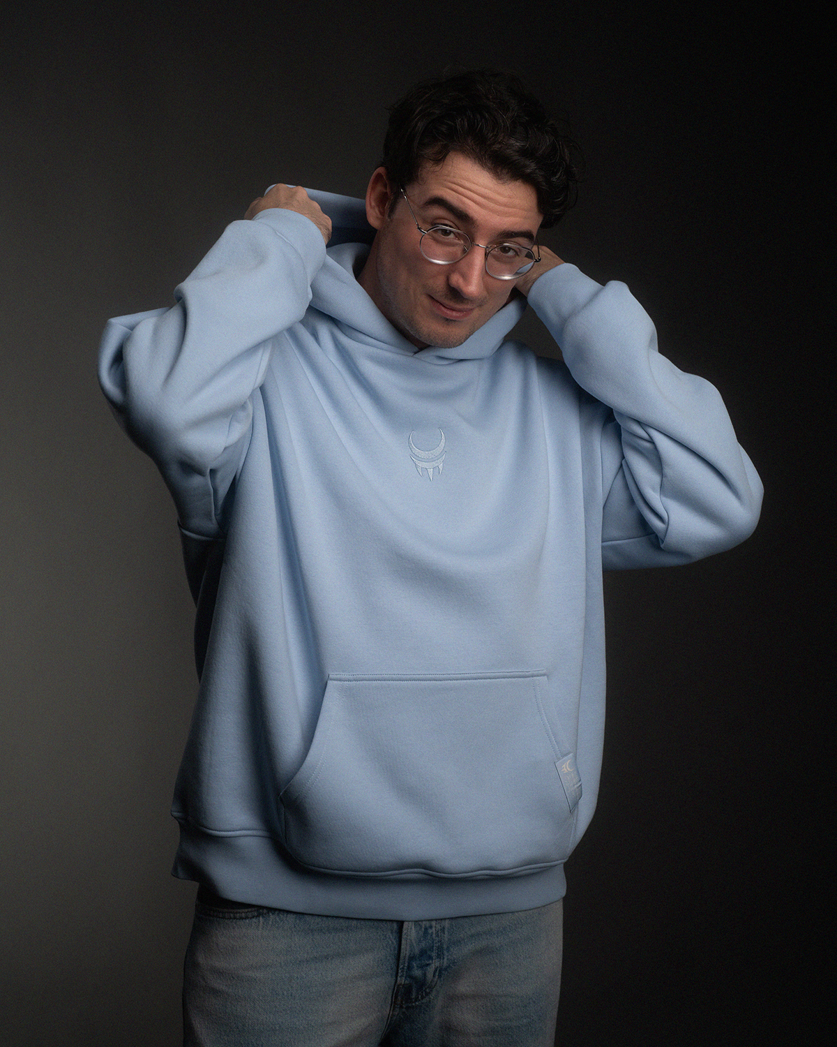 Mann mit hochgezogener Kapuze trägt einen hellblauen Hoodie. Auf der Brust ist ein weißes gesticktes Logo mit Mondkrallen-Symbol zu sehen. Der Hoodie hat eine Kängurutasche.