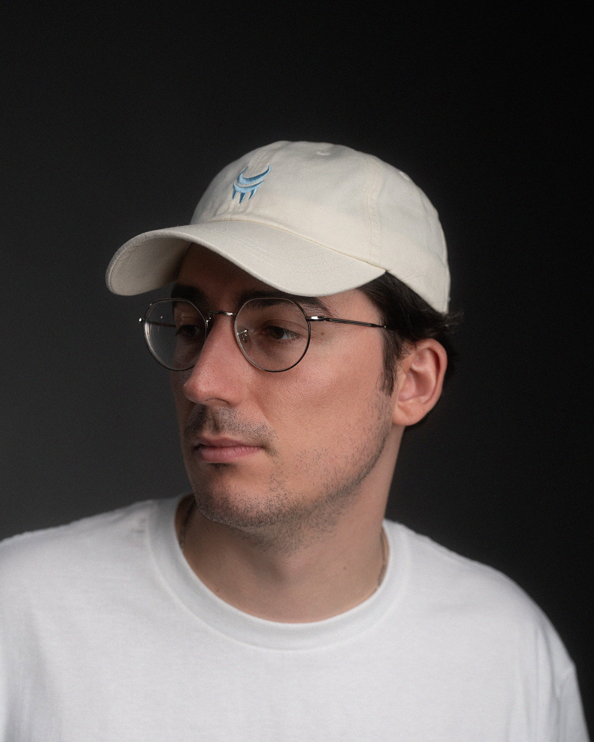 Cremefarbene Baseball-Cap mit hellblauer Stickerei aus Sichel und Krallenmotiv, getragen von einer Person mit Brille und weißem T-Shirt im Halbprofil.
