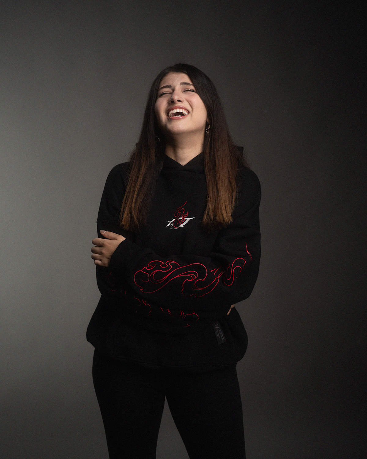 Schwarzer Hoodie mit rotem Flammenmotiv auf Brust und Ärmeln. Die Trägerin lacht mit verschränkten Armen. Fokus auf lebendigen Ausdruck.
