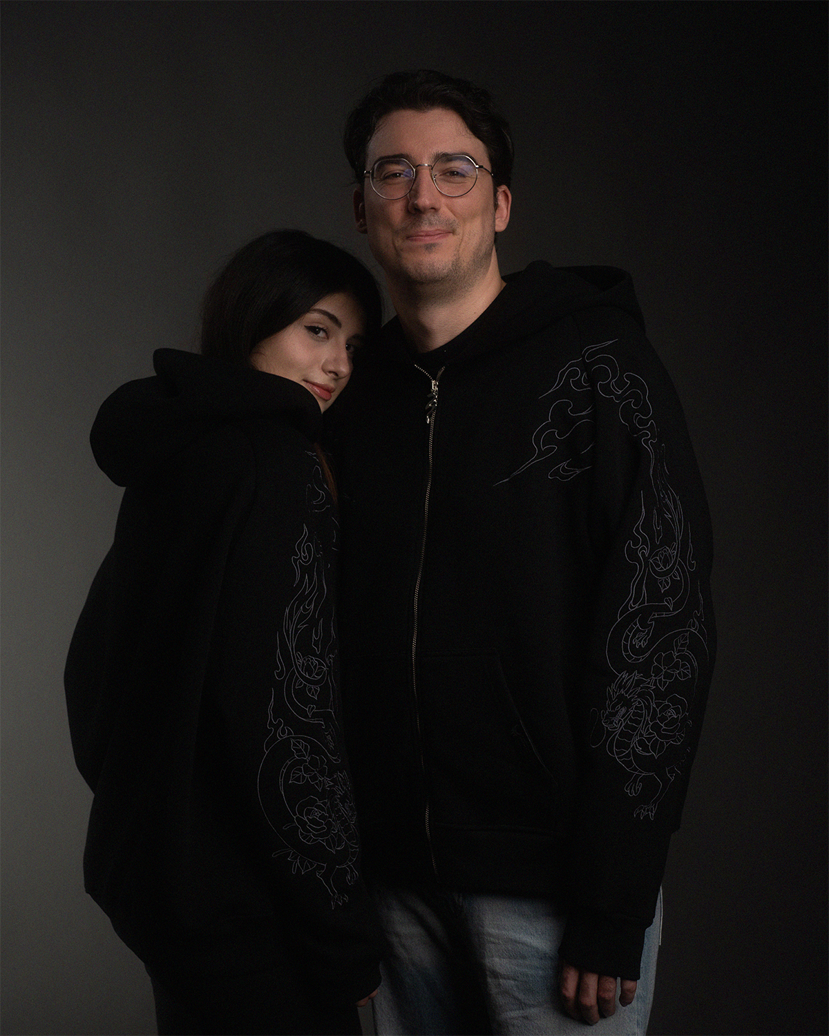 Paaraufnahme: beide Personen tragen die schwarze Kapuzenjacke mit identischer Stickerei. Das Design auf den Ärmeln hebt sich subtil vom Stoff ab.