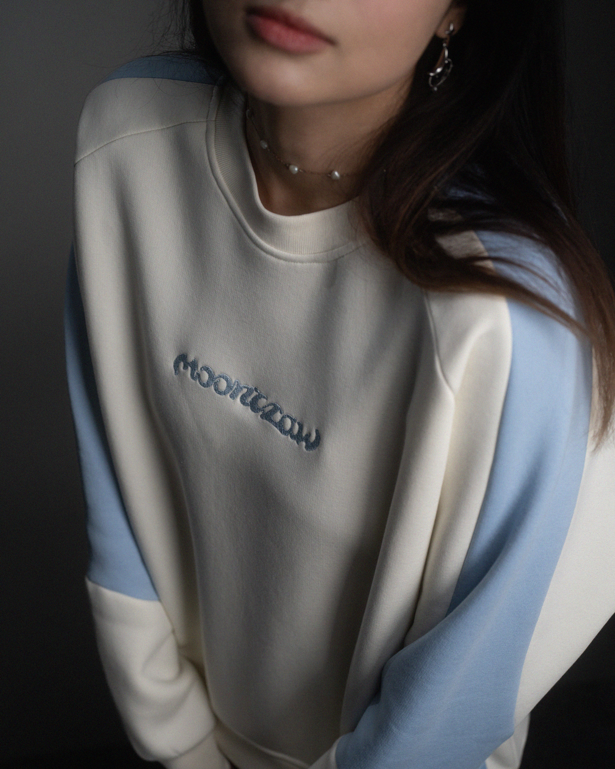 Nahaufnahme eines cremefarbenen Sweatshirts mit hellblauen Ärmeln. Im Zentrum des Bildes ist der hellblau gestickte Schriftzug „moonclaw“ erkennbar.