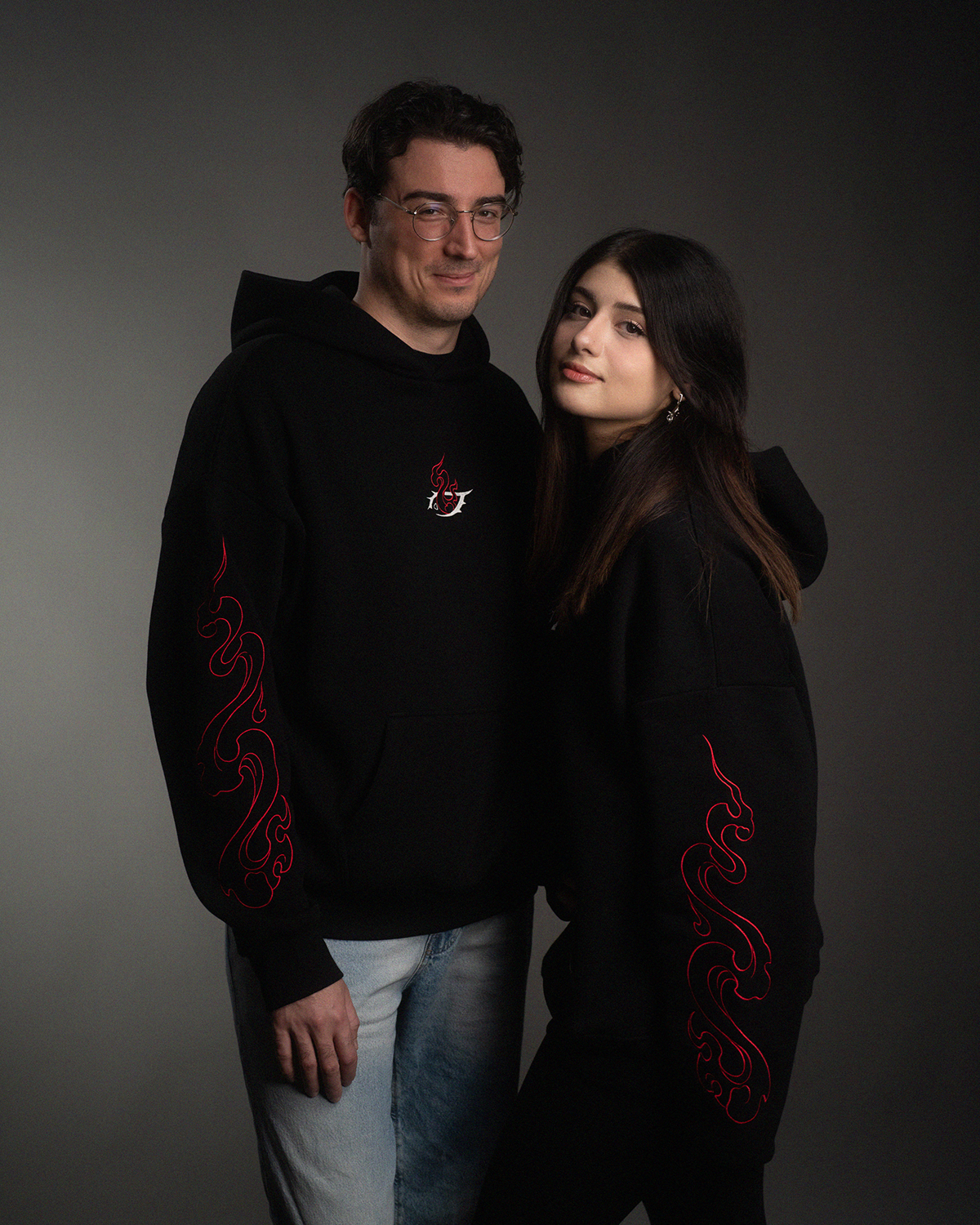 Zwei Personen tragen denselben schwarzen Hoodie mit zentralem Stickmotiv und roten Flammenmustern auf den Ärmeln. Darstellung als Partnerlook.