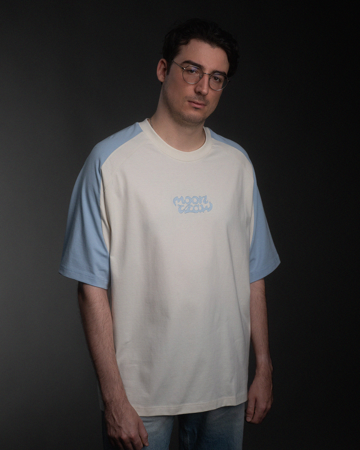 Vorderansicht des Off-White-Shirts mit hellblauen Raglanärmeln und hellblauer „moonclaw“-Stickerei auf der Brust. Das Shirt hat einen lockeren Unisex-Schnitt.