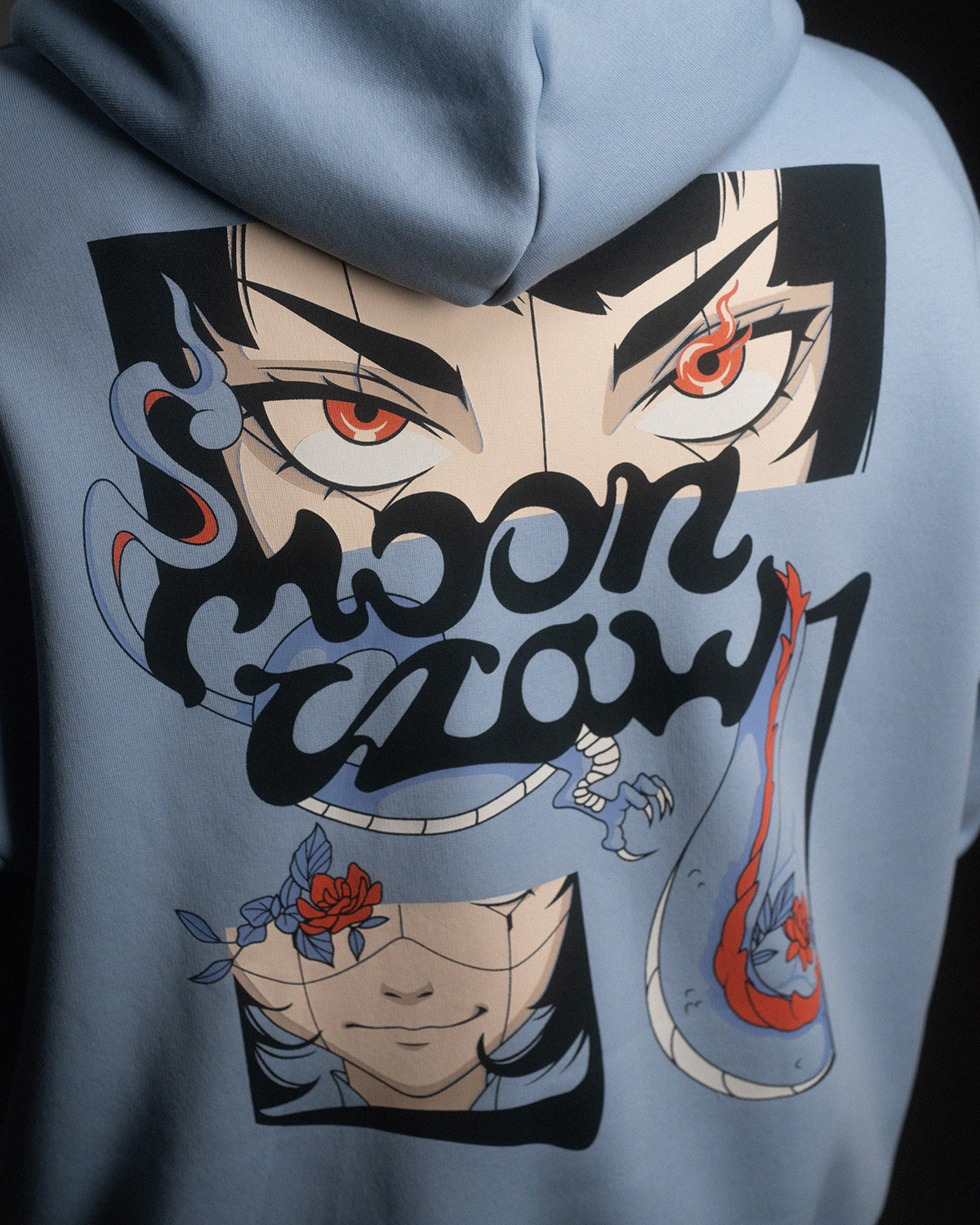 Nahaufnahme des Rückenprints auf hellblauem Hoodie. Der bunte Druck zeigt ein gespiegeltes „moon claw“-Logo, zwei Gesichter, Blüten und flammenartige Formen in kontrastreichen Farben.
