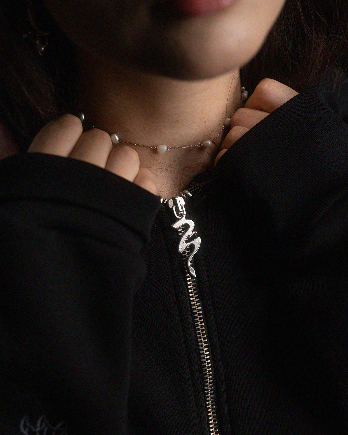 Nahaufnahme des Reißverschlusses der schwarzen Kapuzenjacke. Der Zipper zeigt ein silbernes Metallelement in Form einer stilisierten Schlange.