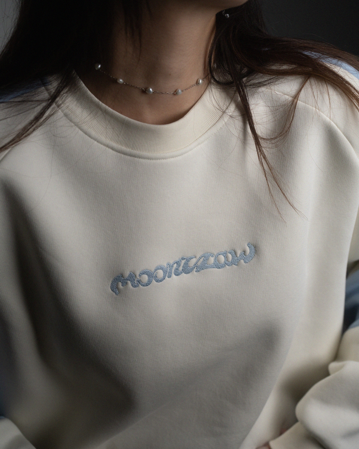 Nahaufnahme des Schriftzugs „moonclaw“ in hellblauer Stickerei auf einem cremefarbenen Sweatshirt mit hellblauen Ärmeln.