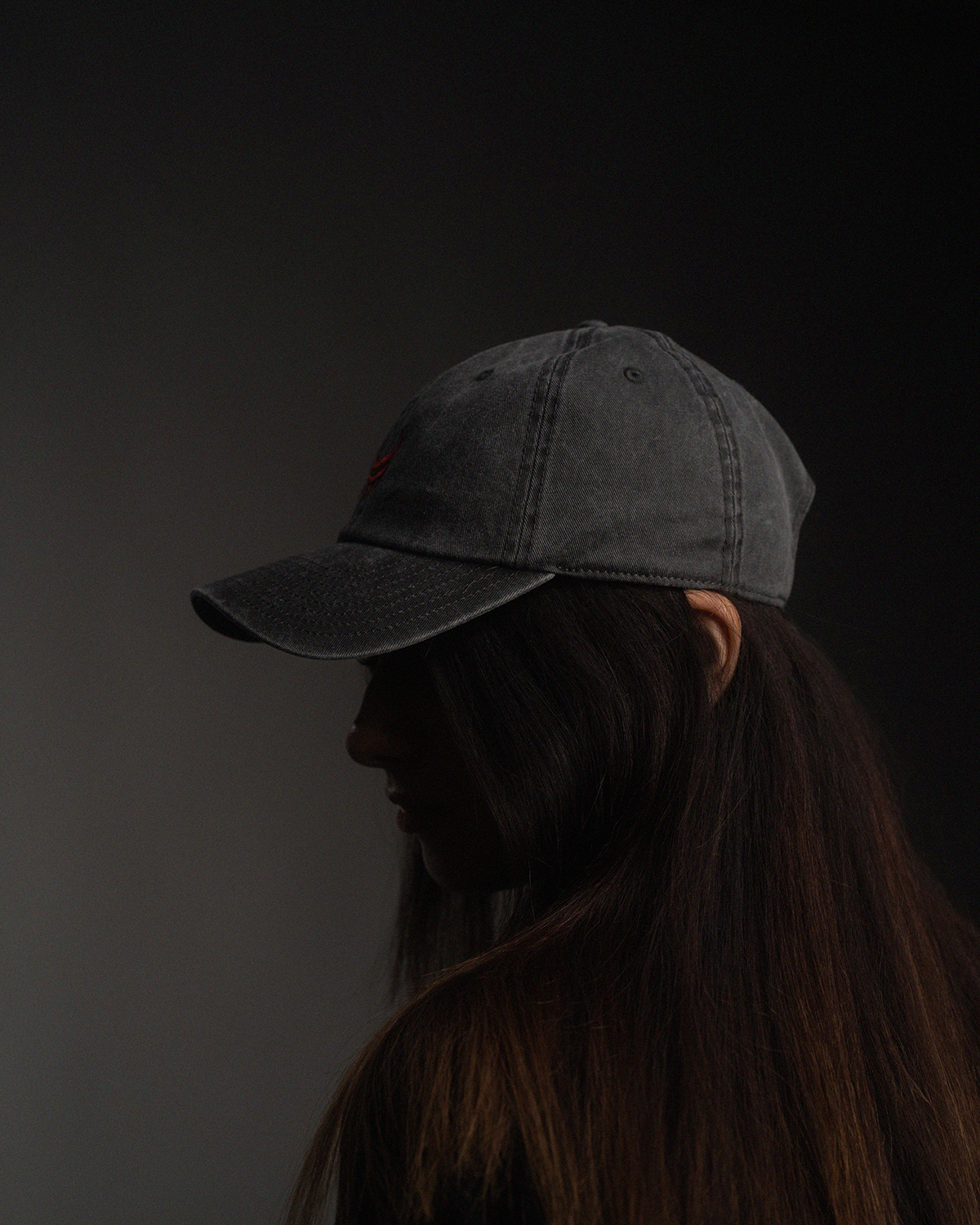Seitlich aufgenommene Cap in verwaschenem Schwarz mit roter Stickerei. Die Cap wird von einer Person mit offenem Haar getragen, das den Rücken bedeckt.