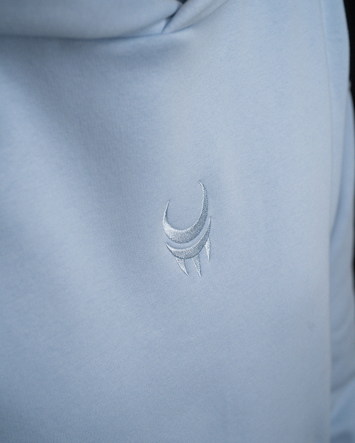 Nahaufnahme des gestickten Logos auf der Brust eines hellblauen Hoodies. Das Motiv ist tonal in Weiß und zeigt ein stilisiertes Mondkrallen-Symbol.