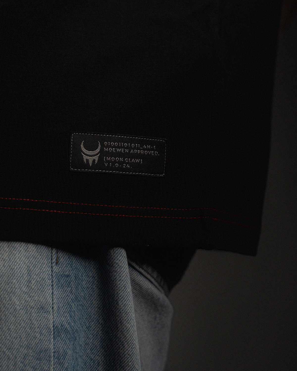 Detailansicht des gewebten Labels am Saum des schwarzen Shirts, mit weißem Mondkrallen-Logo und technischem Textaufdruck.