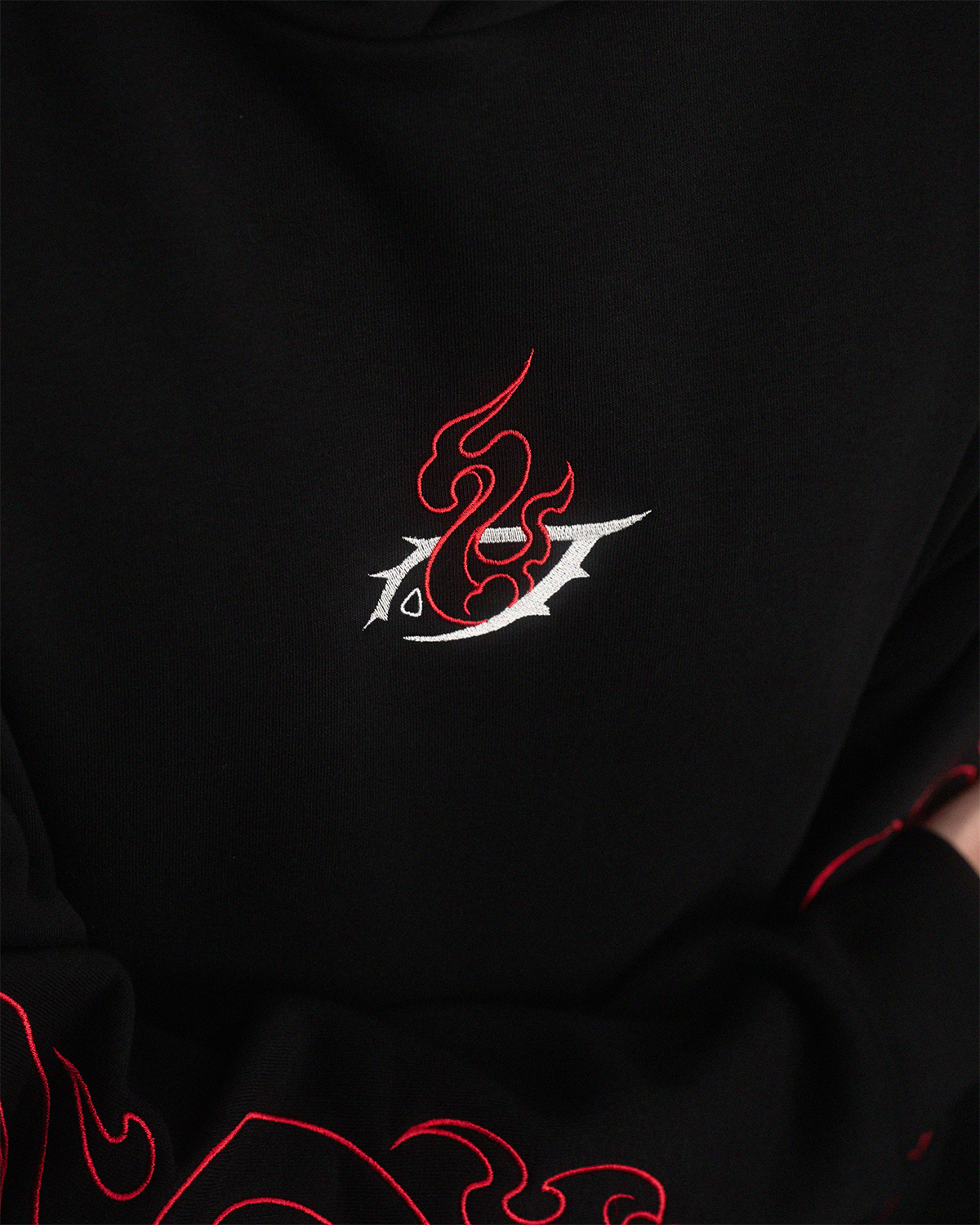 Nahaufnahme des zentralen Brustmotivs auf schwarzem Hoodie, bestehend aus roter Flamme und weißem Symbol. Klare Sicht auf die Stickqualität.