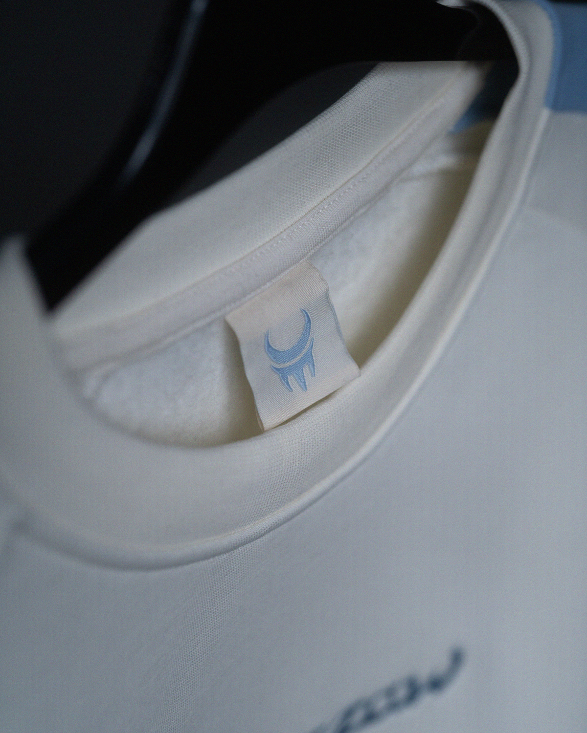 Nahaufnahme des Nackenbereichs eines cremefarbenen Sweatshirts mit hellblauem Webetikett. Auf dem Etikett ist ein stilisiertes Mondsichel-Logo mit Zacken in Hellblau zu sehen.