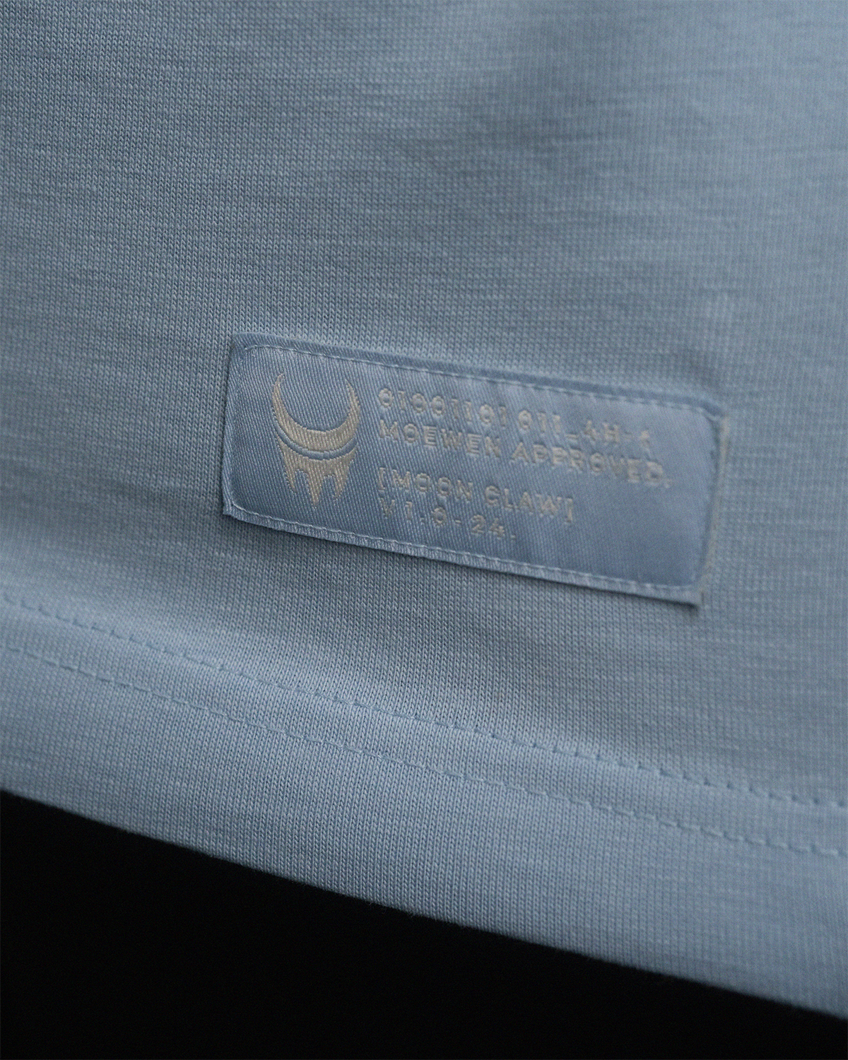 Detailaufnahme des gewebten Labels am unteren Saum eines hellblauen T-Shirts mit „MoonClaw“-Logo und Seriennummer.