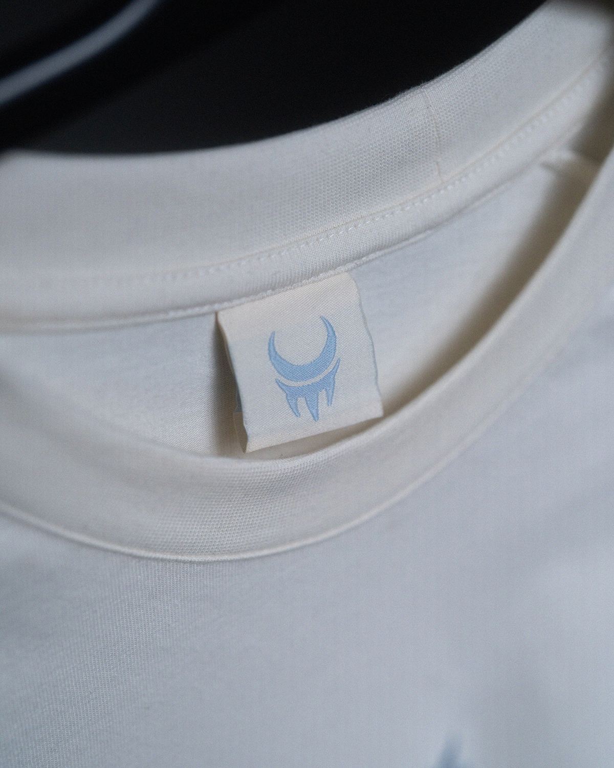 Detailaufnahme des Nackenlabels im Shirt. Das gewebte Etikett zeigt ein hellblaues Mondkrallen-Logo auf weißem Hintergrund.