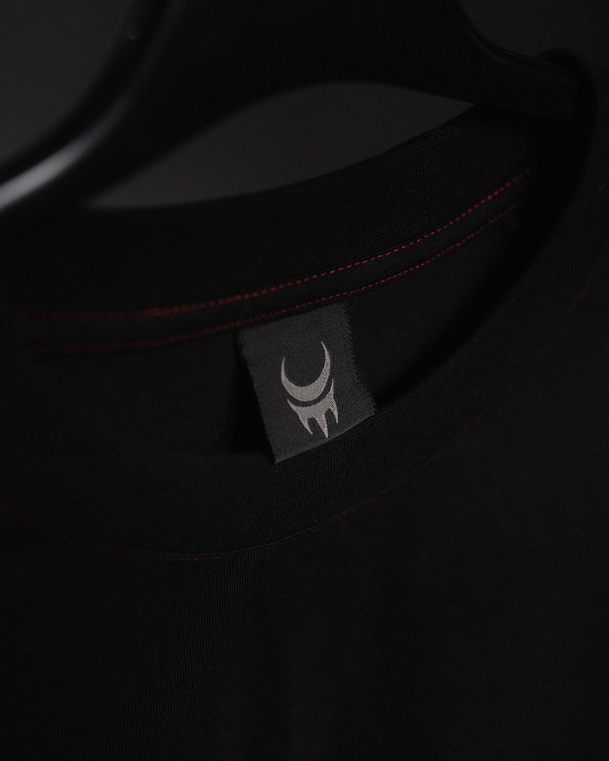 Nahaufnahme des Nackenbereichs eines schwarzen T-Shirts auf einem Kleiderbügel. Sichtbar ist ein graues Webetikett mit dem hellen Mondkrallen-Logo der Marke MoonClaw. Der Rundhalsausschnitt ist mit einer auffälligen roten Ziernaht versehen.