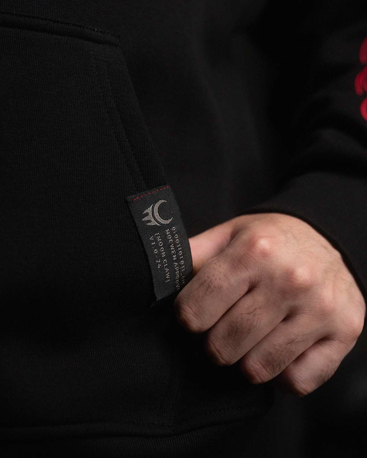 Detailansicht des schwarzen Labels mit „Moon Claw“-Symbol und technischer Aufschrift, platziert an der Seitentasche des Hoodies.