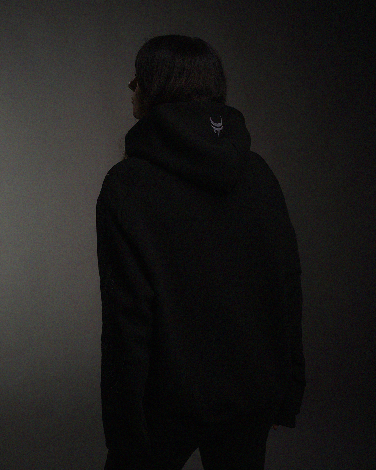 Die Jacke wird von hinten gezeigt, getragen von einer Person mit langen Haaren. Der Fokus liegt auf dem gestickten Logo auf der Kapuze.