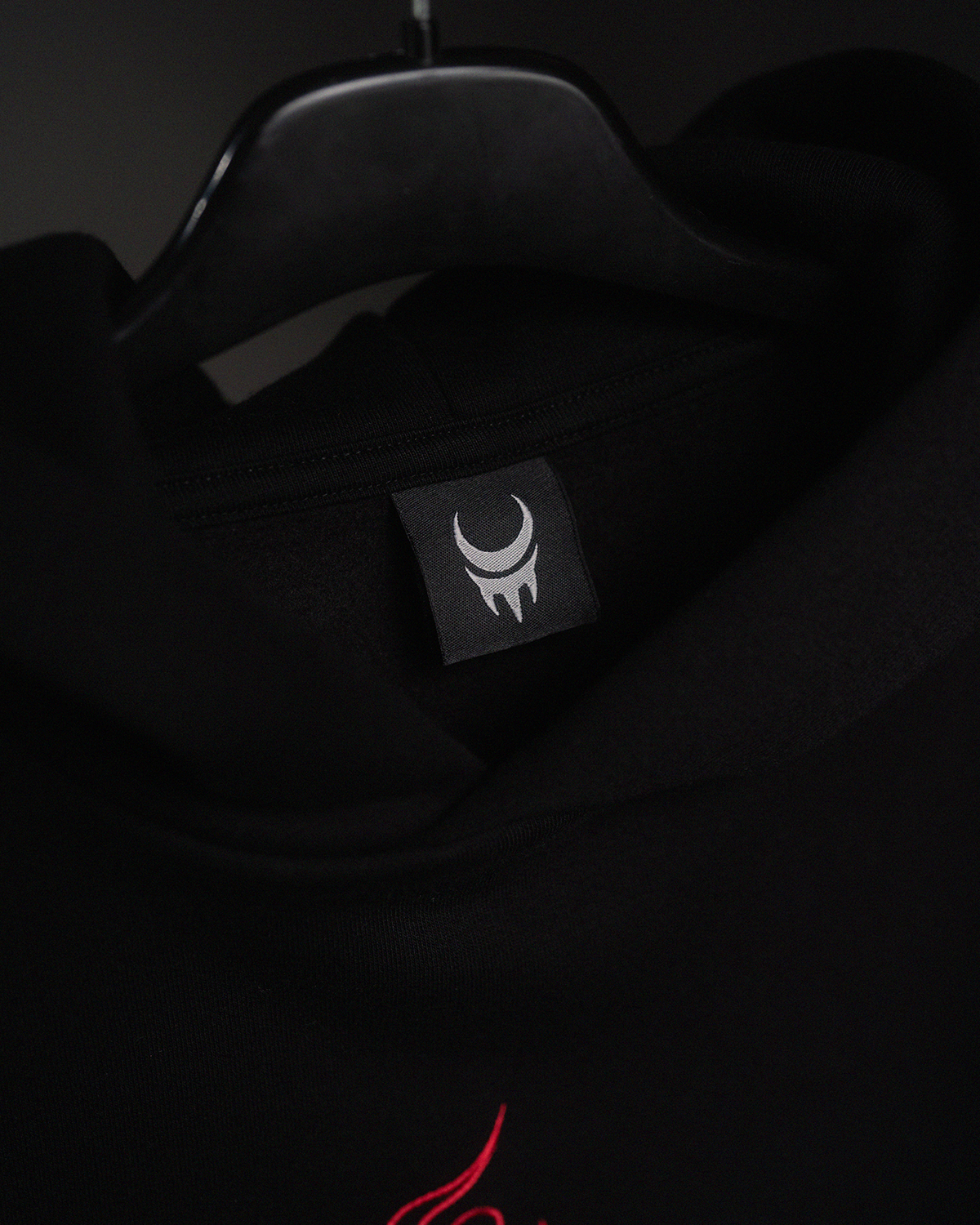 Nahaufnahme des schwarzen Nackenlabels mit weißem Sichelmondsymbol in Krallenform auf schwarzem Hoodie. Kleidungsstück hängt auf Bügel.