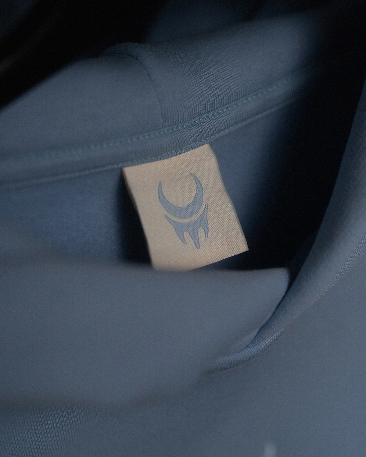 Etikett im Nacken eines hellblauen Hoodies mit aufgedrucktem „moon claw“-Symbol in blassem Blau auf beigefarbenem Stoff.