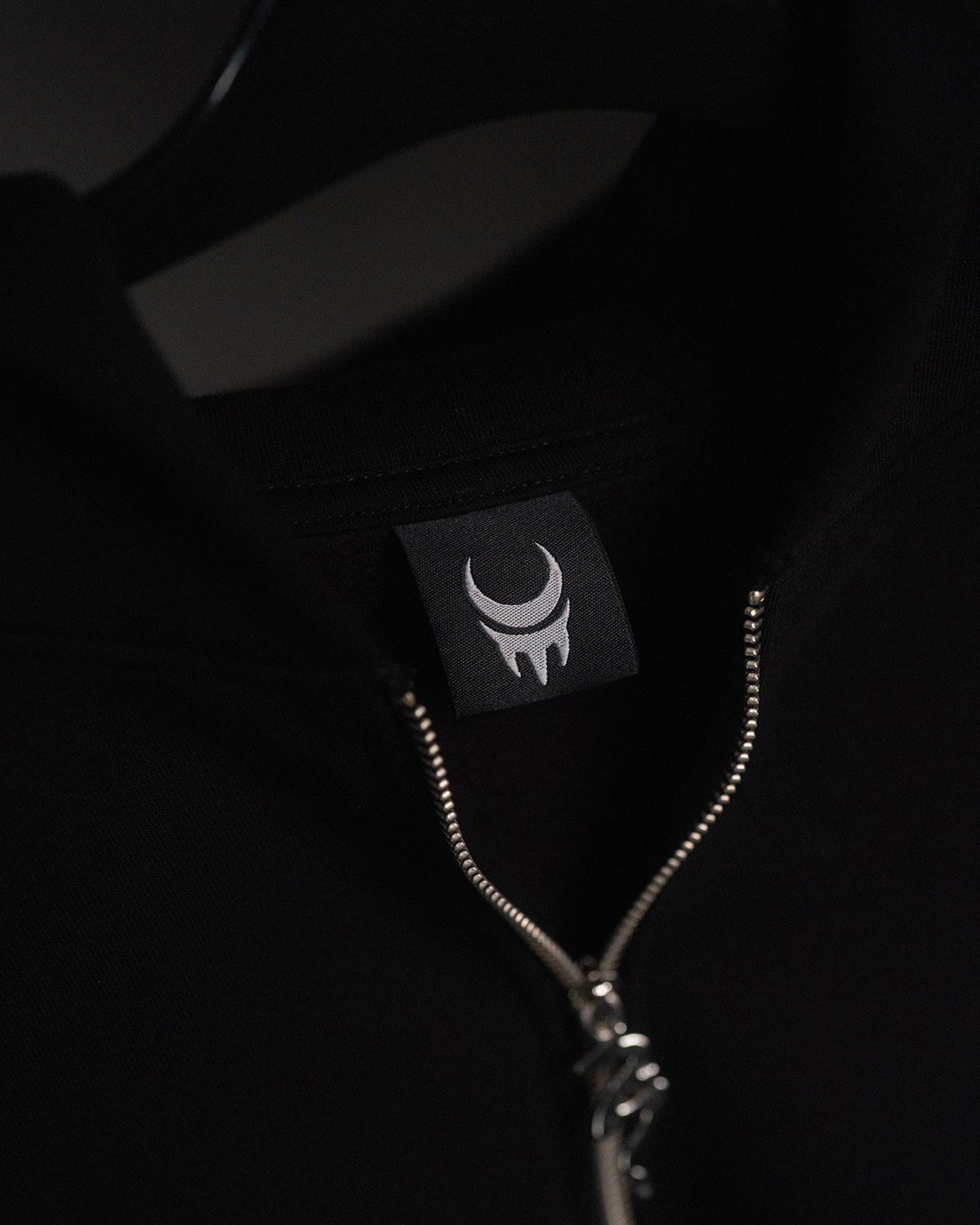 Detailaufnahme des Nackenetiketts der Jacke, das ein helles Mondsichel-Logo auf schwarzem Stoff zeigt. Der Reißverschluss ist leicht geöffnet.