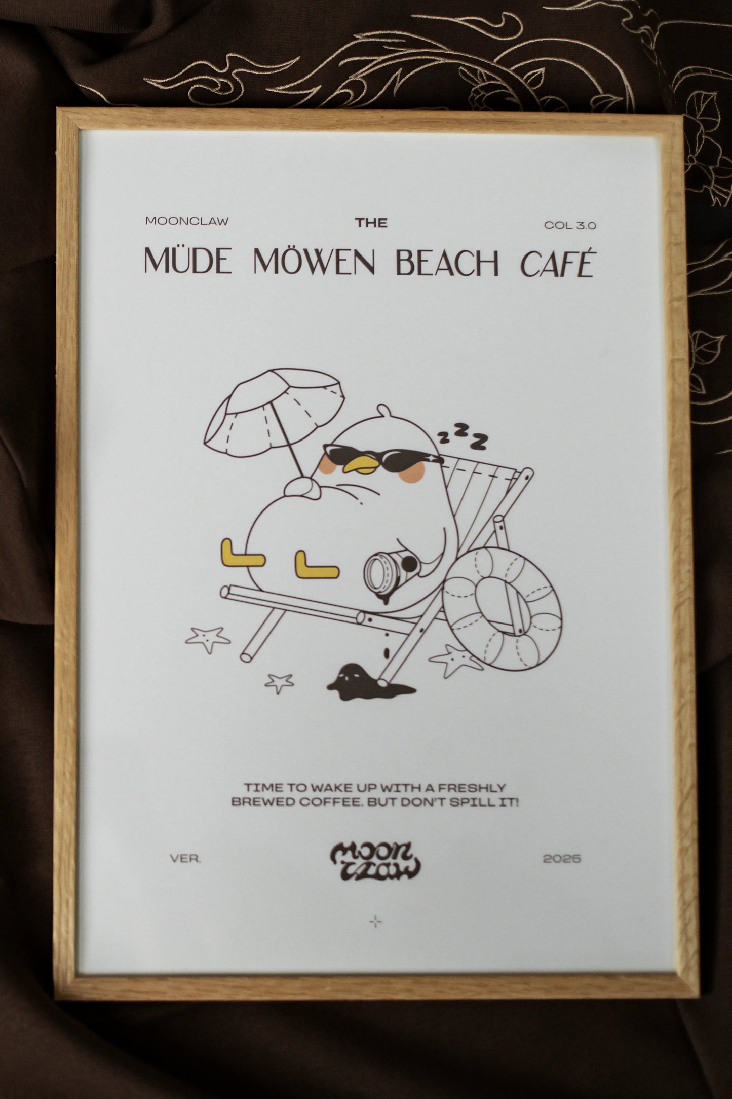 Gerahmtes Poster mit der Aufschrift „MÜDE MÖWEN BEACH CAFÉ“ und Illustration einer entspannt liegenden Möwe mit Sonnenbrille im Liegestuhl unter Sonnenschirm. Unter dem Motiv steht der Satz „Time to wake up with a freshly brewed coffee. But don’t spill it!“. Der Druck ist in einem hellen Holzrahmen gefasst und liegt auf braunem Stoff mit feinen Stickmustern.