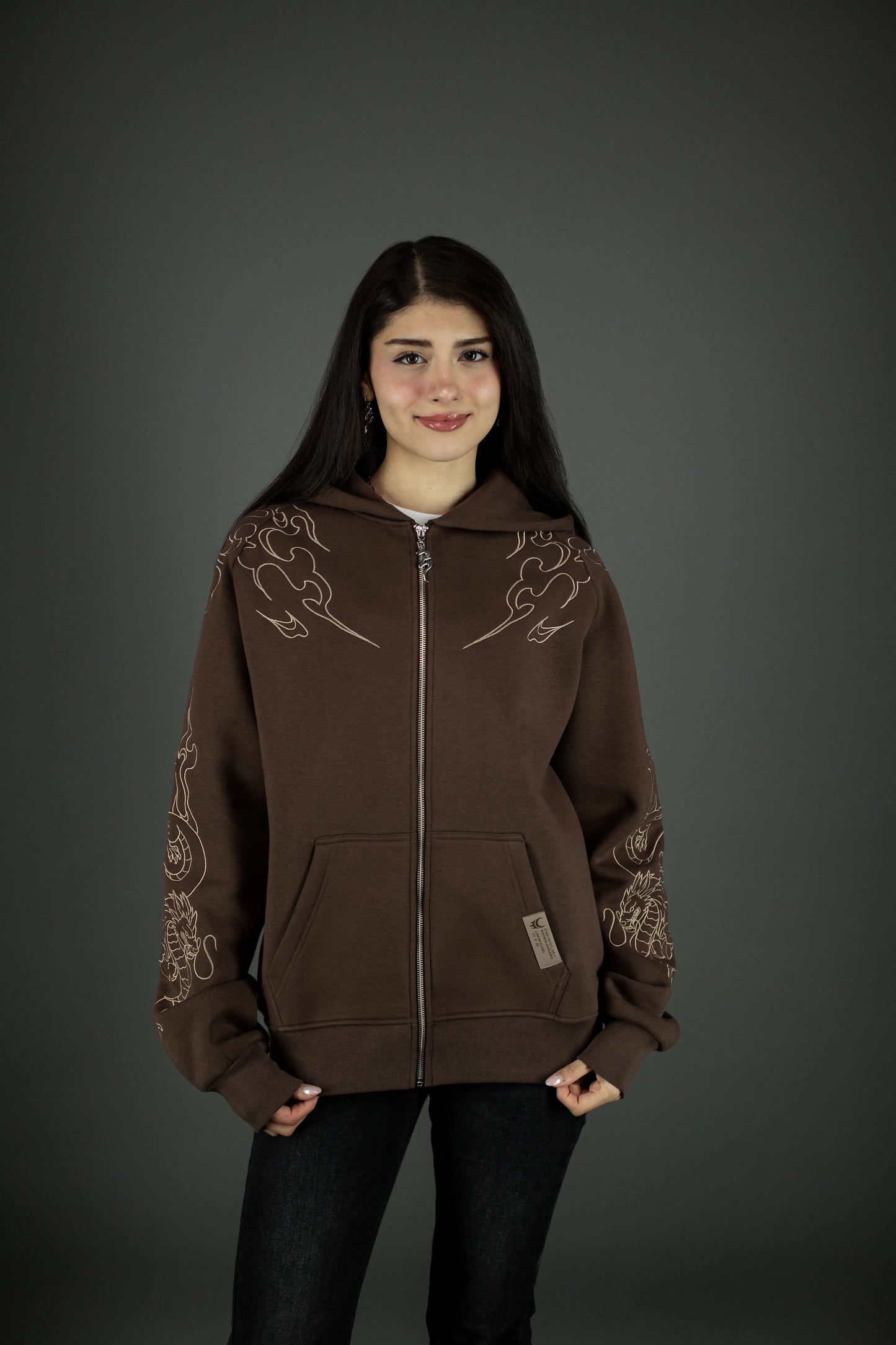 Brauner Zip-Hoodie mit beigem Flammendesign auf den Schultern und Drachen-Stickerei an den Ärmeln. Der Hoodie wird geschlossen getragen, mit sichtbarer Fronttasche und kleinem Markenlabel am Saum.