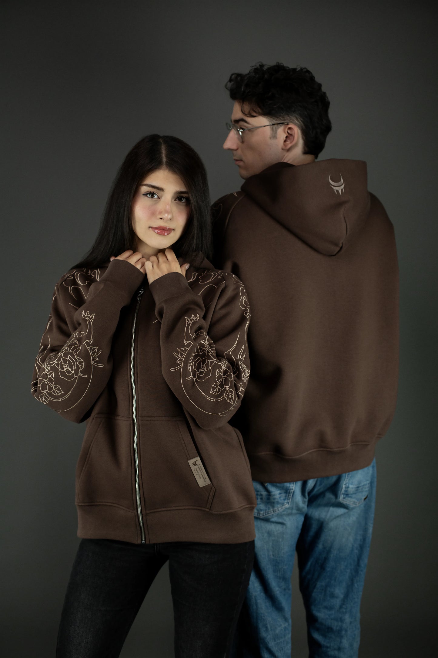 Zwei Personen tragen den braunen Hoodie. Die vordere Person zeigt die Front mit Drachenmuster an den Ärmeln, die hintere Person steht mit dem Rücken zur Kamera und zeigt das gestickte Mondsymbol auf der Kapuze.