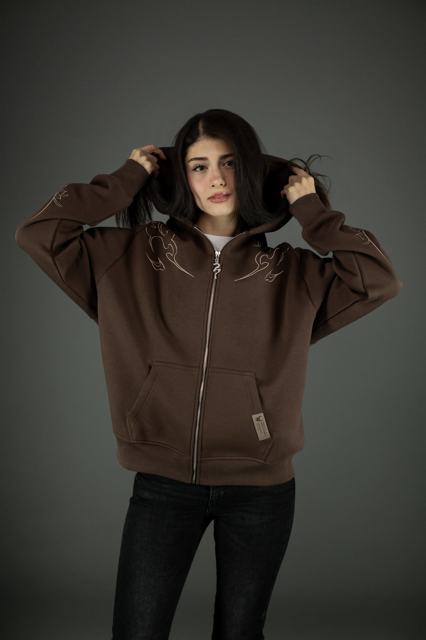 Frontansicht eines braunen Zip-Hoodies mit beigefarbenem Flammenmuster über den Schultern. Die Kapuze ist aufgesetzt, der Reißverschluss geschlossen, der Stoff wirkt dick und strukturiert.