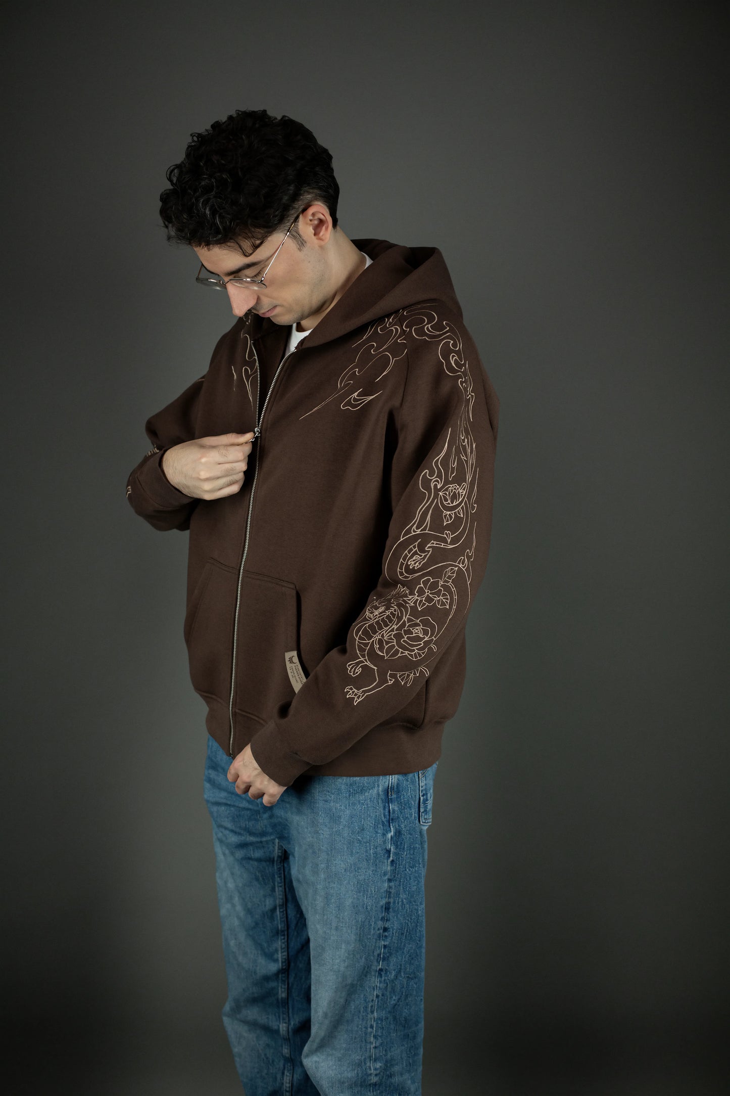 Der Hoodie wird von einer Person getragen, die den metallischen Reißverschluss schließt. Auf dem Ärmel ist das florale Drachenmotiv sichtbar, das sich von der Schulter bis zum Handgelenk zieht.