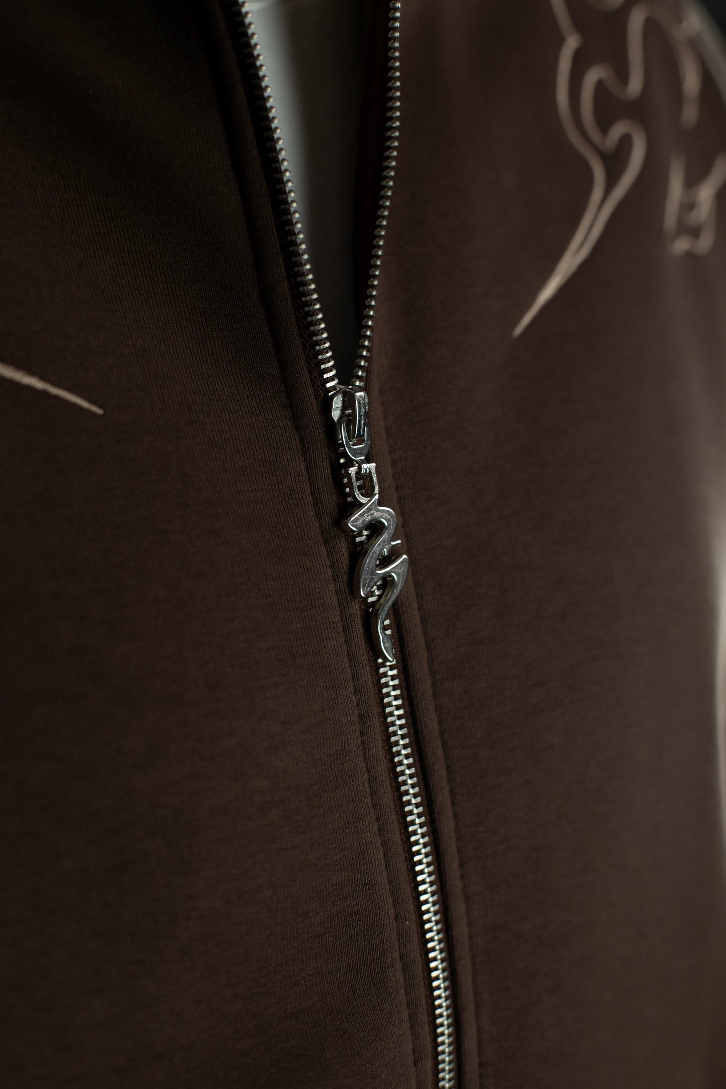 Detail des silbernen Reißverschlusses des Hoodies, dessen Zipper in Form einer geschwungenen Drachenfigur gestaltet ist. Der Stoff ist in dichter, glatter Struktur sichtbar.