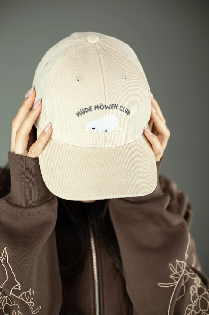 Beigefarbene Cord-Cap mit gesticktem Motiv auf der Vorderseite. Die Stickerei zeigt eine schlafende Möwe mit geschlossenen Augen und gelbem Schnabel, darüber der dunkelgraue Schriftzug „MÜDE MÖWEN CLUB“. Die Cap wird von einer Person getragen, deren Hände den Schirm halten, während der Rest des Gesichts verdeckt ist. Der Stoff der Cap ist fein gerippt und wirkt weich strukturiert.