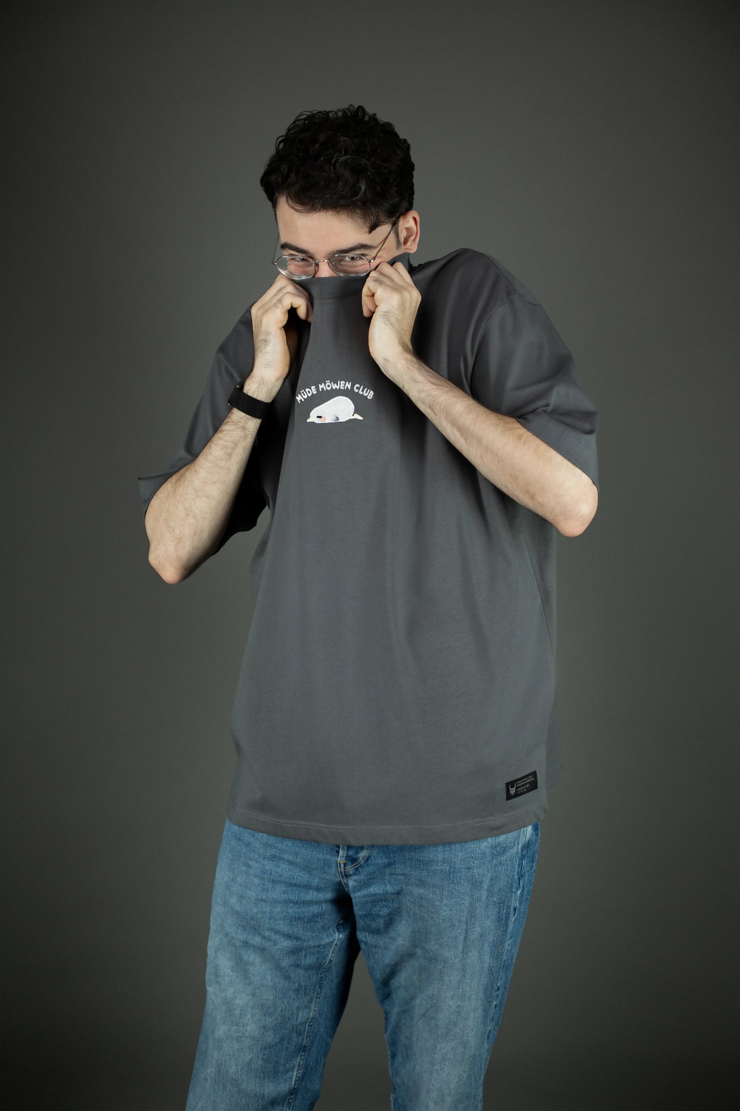 Person trägt das graue T-Shirt mit „MÜDE MÖWEN CLUB“-Stickerei und zieht den Stoff spielerisch bis zur Nase. Die Bruststickerei zeigt eine liegende, schlafende Möwe mit hellem Körper und gelbem Schnabel.