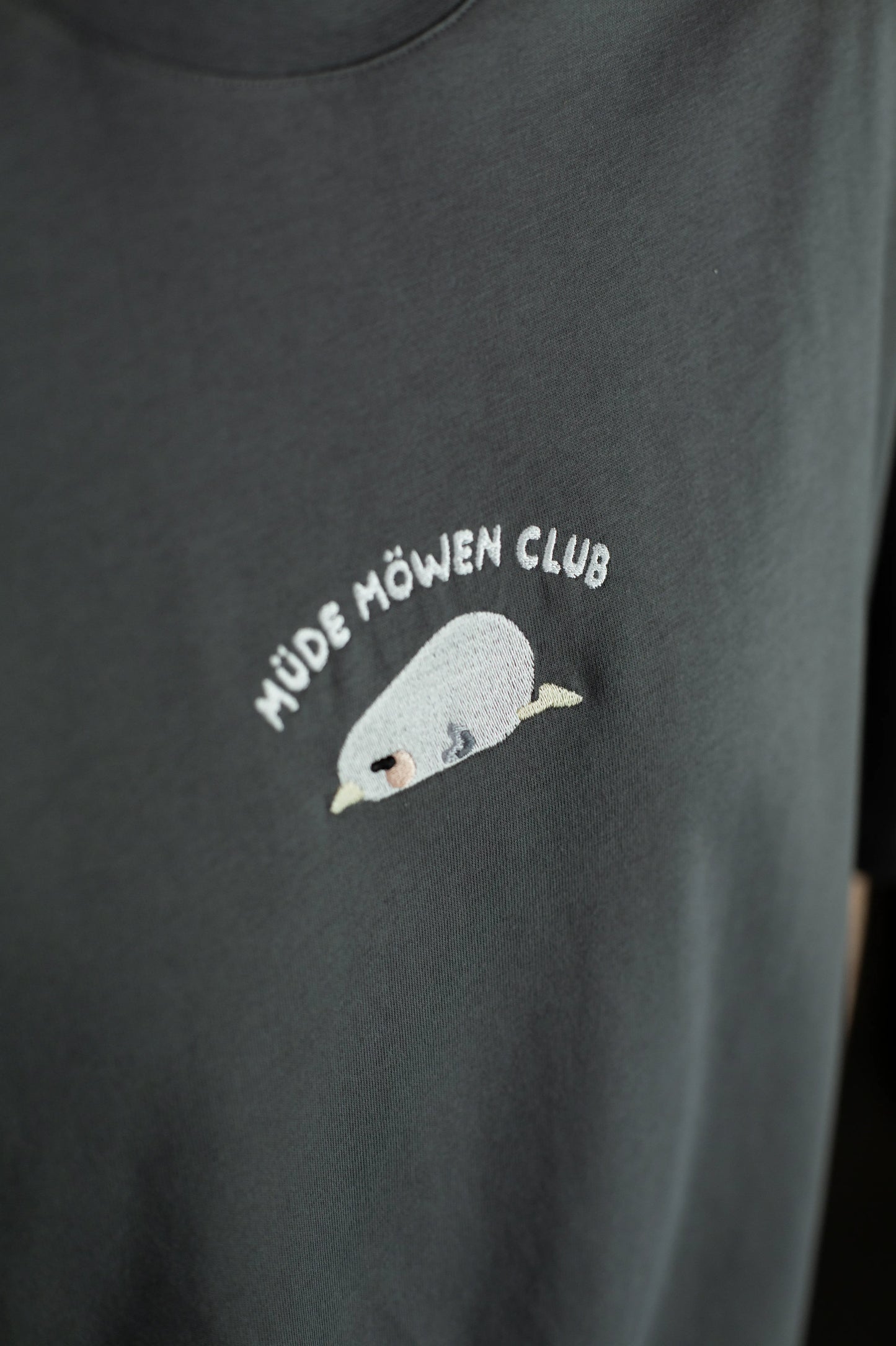 Detailaufnahme der Stickerei auf der Brust: eine schlafende, hellgraue Möwe mit geschlossenem Auge und beigefarbenem Schnabel unter dem Schriftzug „MÜDE MÖWEN CLUB“.