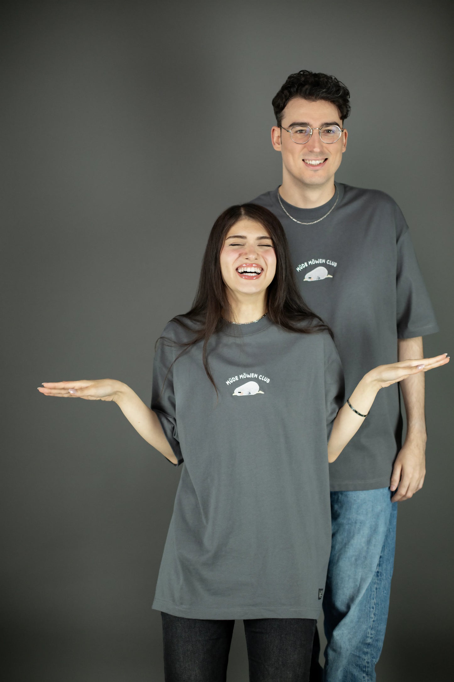 Zwei Personen tragen ein graues T-Shirt mit weißer Stickerei „MÜDE MÖWEN CLUB“ und einer schlafenden Möwe auf der Brust. Das Shirt hat eine weite Passform und kurze Ärmel. Beide lachen in entspannter Pose vor neutralem Hintergrund.