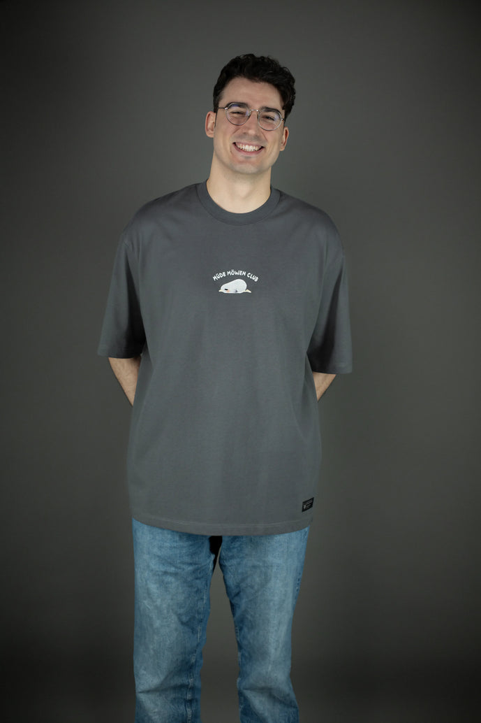 Frontansicht des grauen T-Shirts mit gesticktem Schriftzug „MÜDE MÖWEN CLUB“ über einer liegenden Möwe. Das Shirt ist locker geschnitten und endet auf Hüfthöhe, kombiniert mit einer hellblauen Jeans.