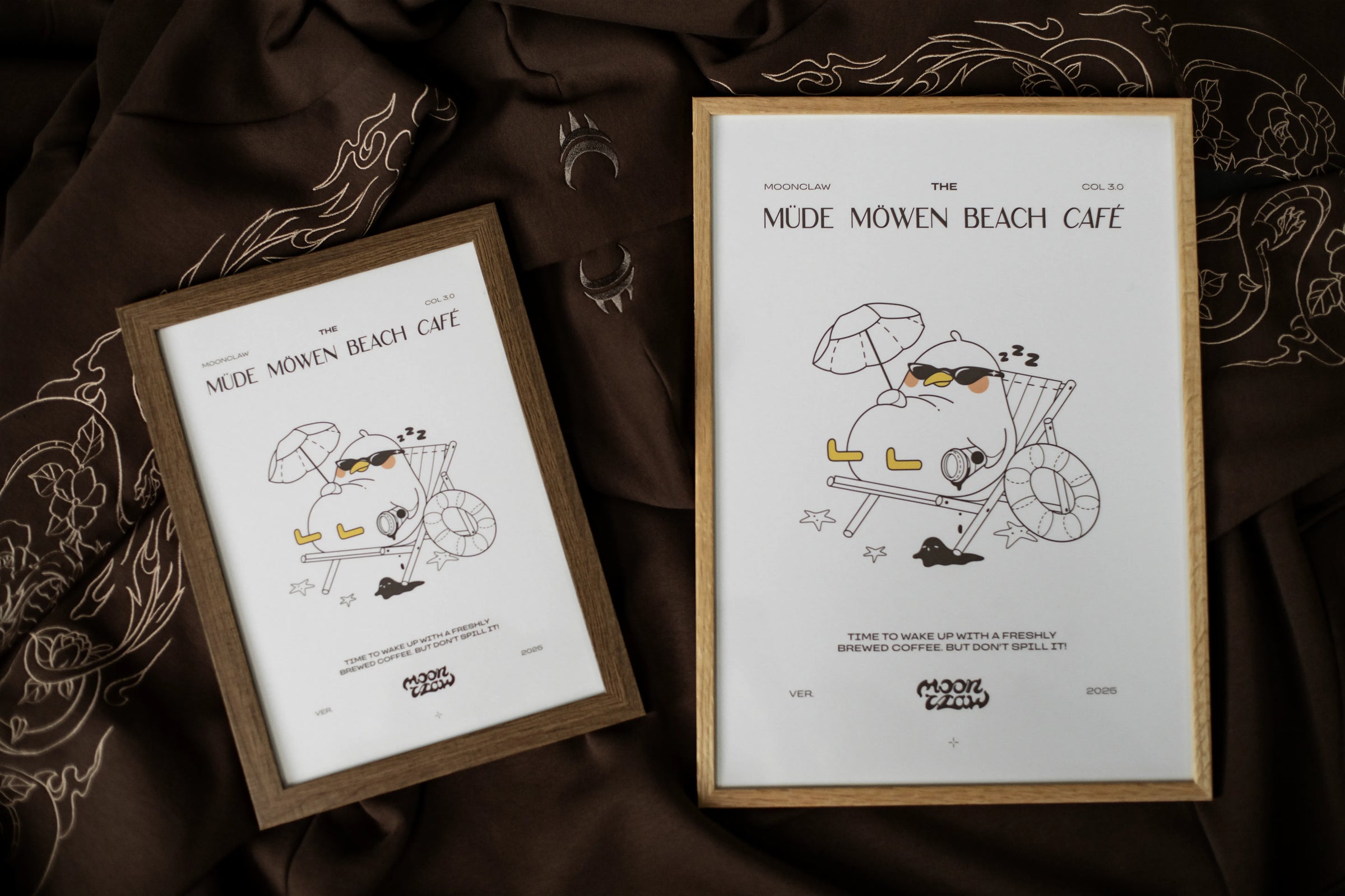 Zwei gerahmte Poster mit identischem Motiv auf braunem Stoffhintergrund. Die Drucke zeigen eine schlafende Möwe mit Sonnenbrille in einem Liegestuhl unter einem Sonnenschirm, daneben der Text „MÜDE MÖWEN BEACH CAFÉ“. Das Motiv ist in einem hellen, minimalistischen Stil gehalten, die Rahmen bestehen aus hellem Holz.