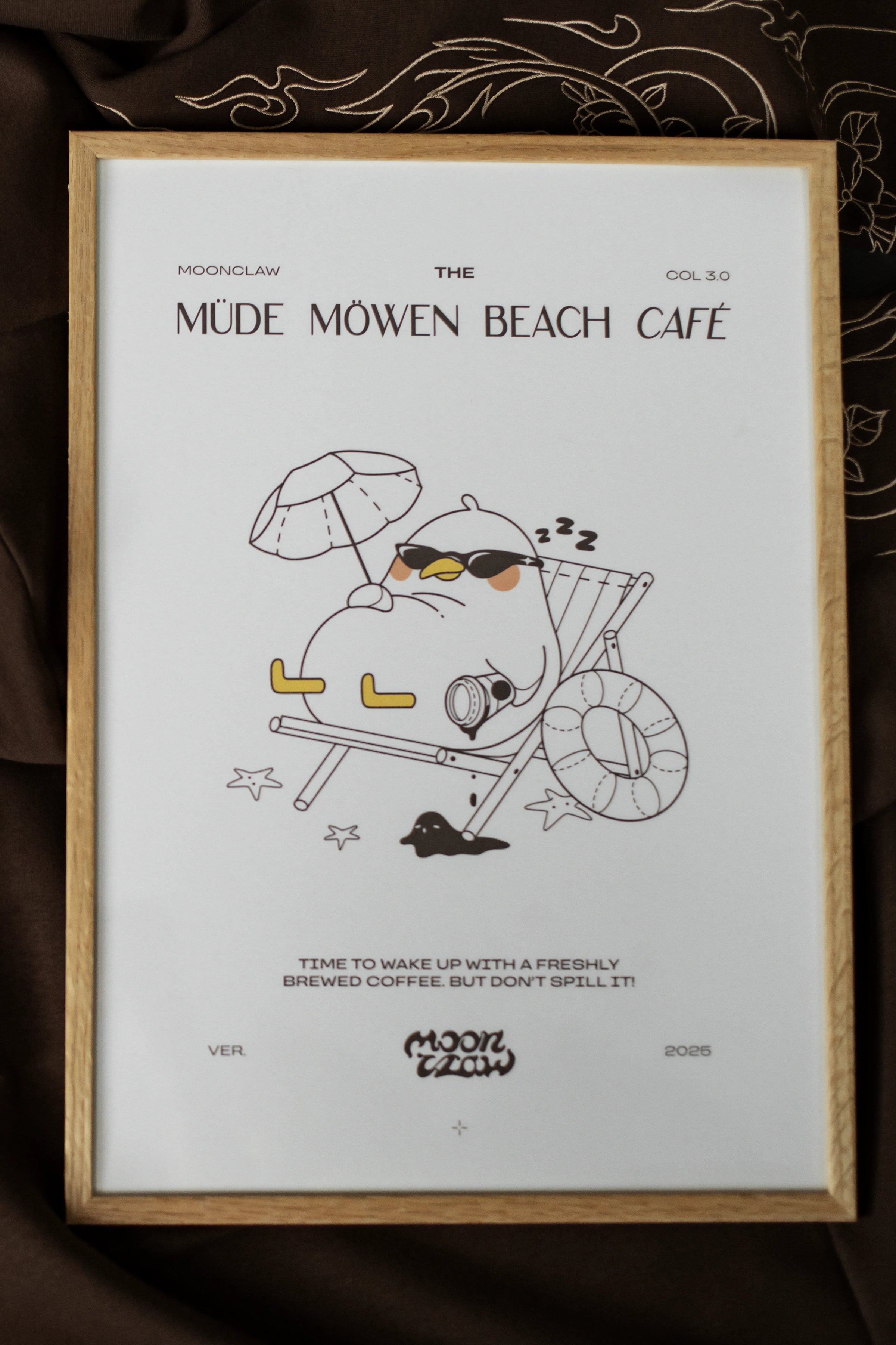 Gerahmtes Poster mit der Aufschrift „MÜDE MÖWEN BEACH CAFÉ“ und Illustration einer entspannt liegenden Möwe mit Sonnenbrille im Liegestuhl unter Sonnenschirm. Unter dem Motiv steht der Satz „Time to wake up with a freshly brewed coffee. But don’t spill it!“. Der Druck ist in einem hellen Holzrahmen gefasst und liegt auf braunem Stoff mit feinen Stickmustern.