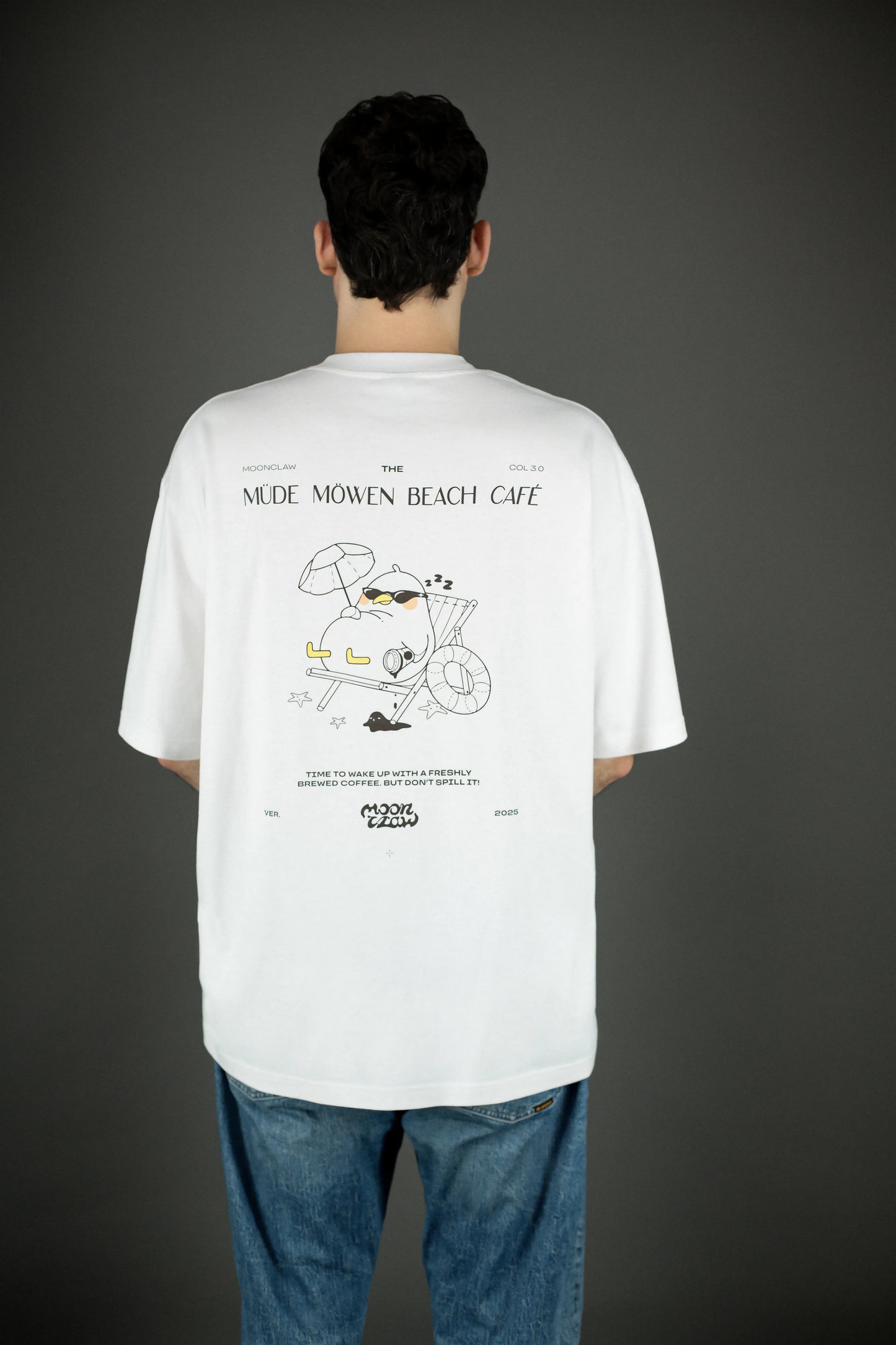 Rückansicht einer Person mit weißem T-Shirt. Der großflächige Rückendruck zeigt die schlafende Möwe mit Sonnenbrille und Kaffeebecher im Liegestuhl unter einem Sonnenschirm, ergänzt durch den Schriftzug „MÜDE MÖWEN BEACH CAFÉ“.