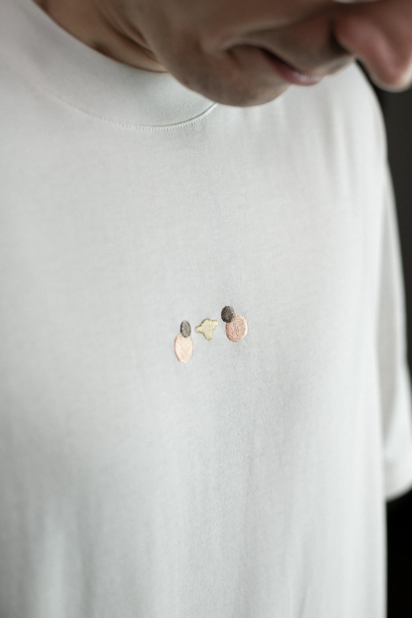 Detailansicht der Vorderseite des weißen T-Shirts mit kleiner, minimalistischer Stickerei in der Mitte der Brust. Die Stickerei zeigt drei kleine Kreise in Beige, Grau und Gelb – eine abstrahierte Darstellung der Möwe aus der Kollektion.